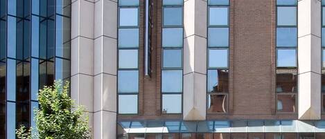 A modern hotel with a prominent 'NH' sign on top, featuring a mix of glass and brick facades, and a clear entrance with a sign reading 'NH PIRINEOS'.
