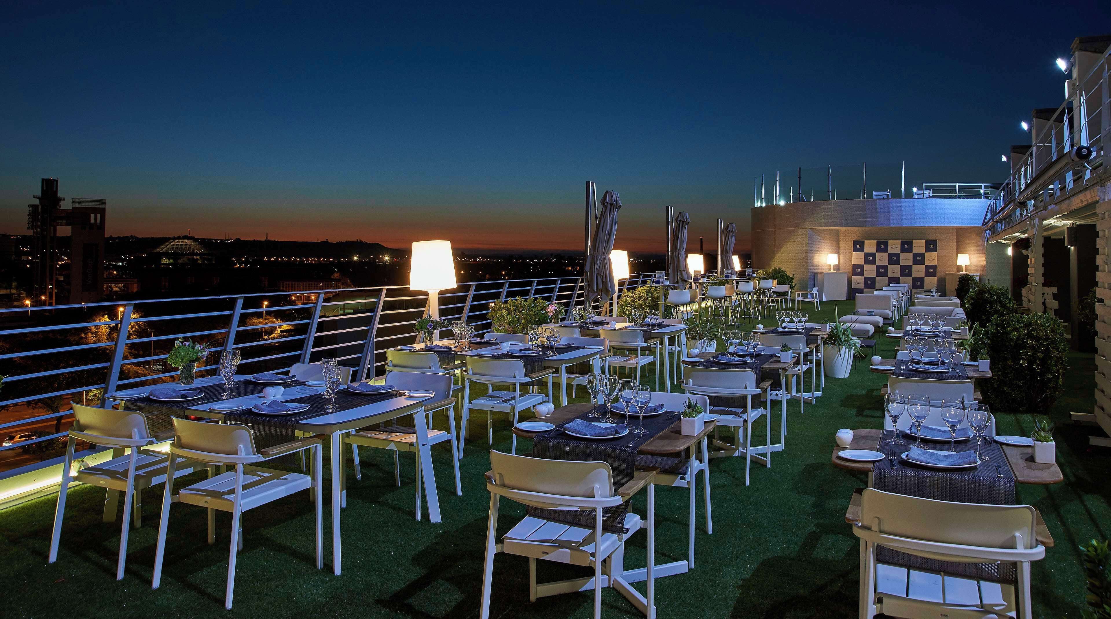 Un área de comedor al aire libre con mesas dispuestas para varios invitados, rodeada por una barandilla con vista panorámica de la ciudad.