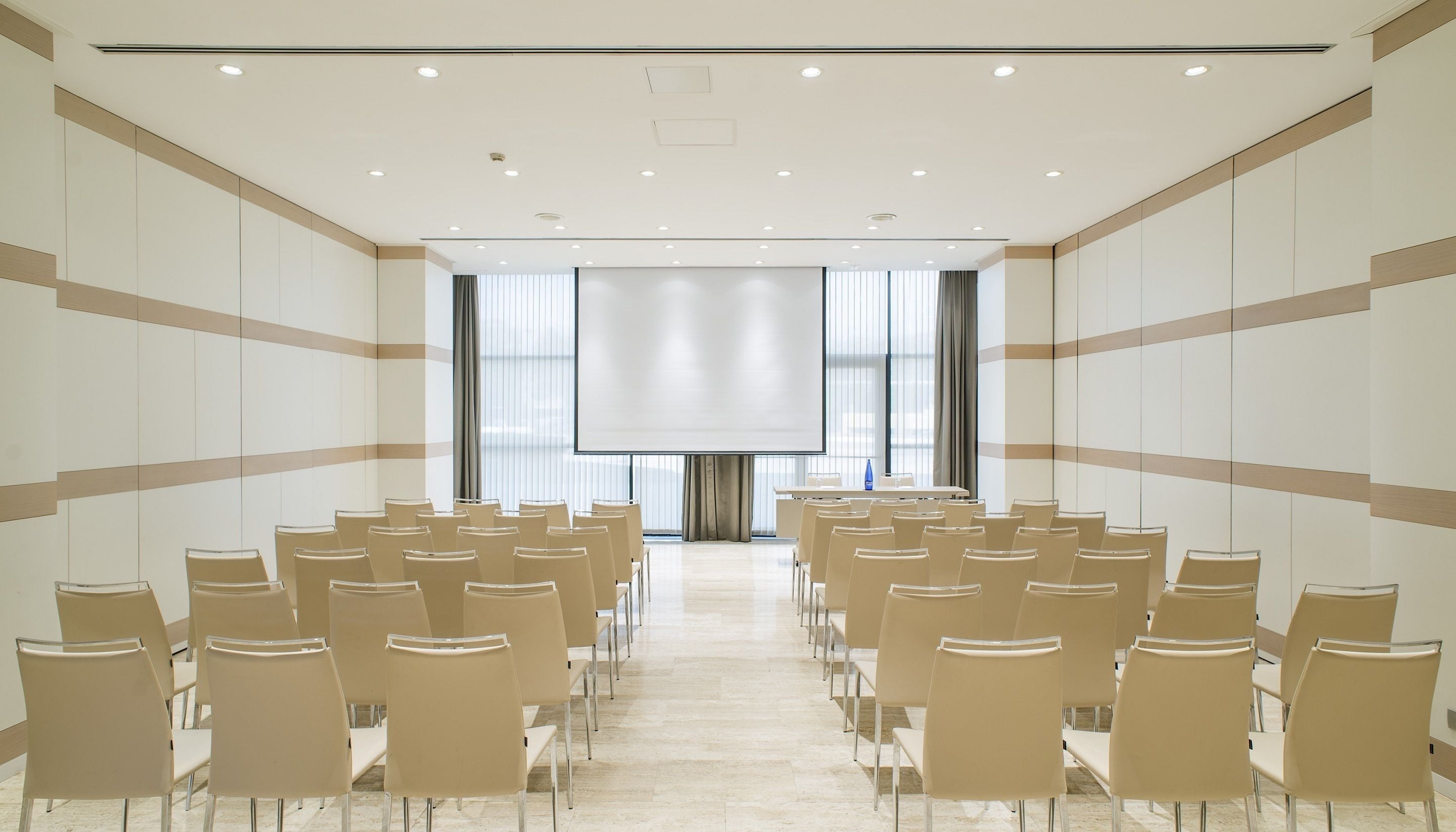 Un salón de conferencias con filas de sillas, una pantalla grande y una mesa larga al frente.