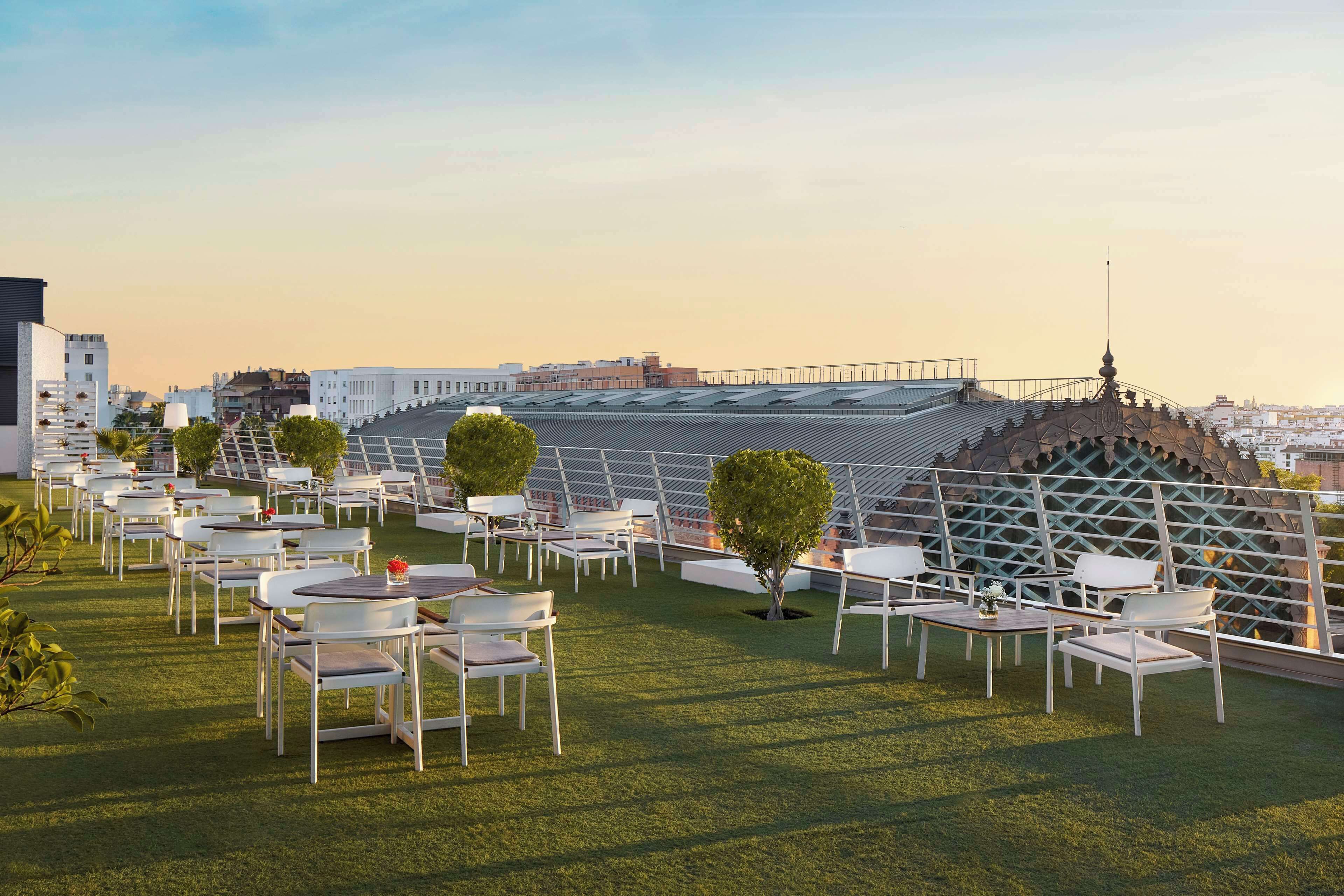 An outdoor seating area with white tables and chairs, a green lawn, and a modern building with a unique roof structure in the background.