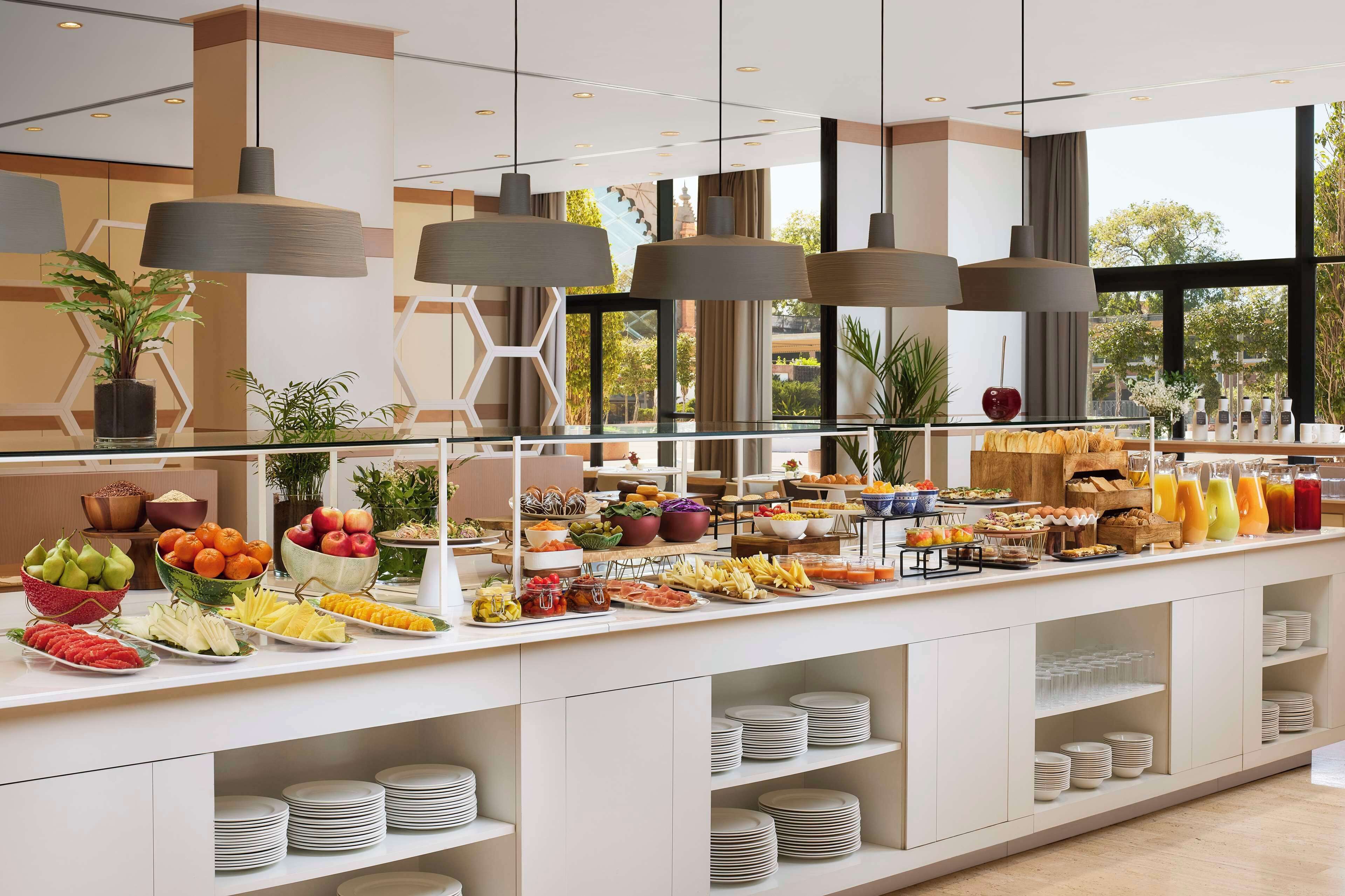 Un bufé con diferentes alimentos y bebidas, un ventanal con vistas y luminarias colgantes.