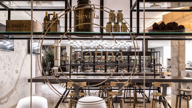 A bar area with shelves displaying bottles and jars, wooden chairs, and a marble wall.