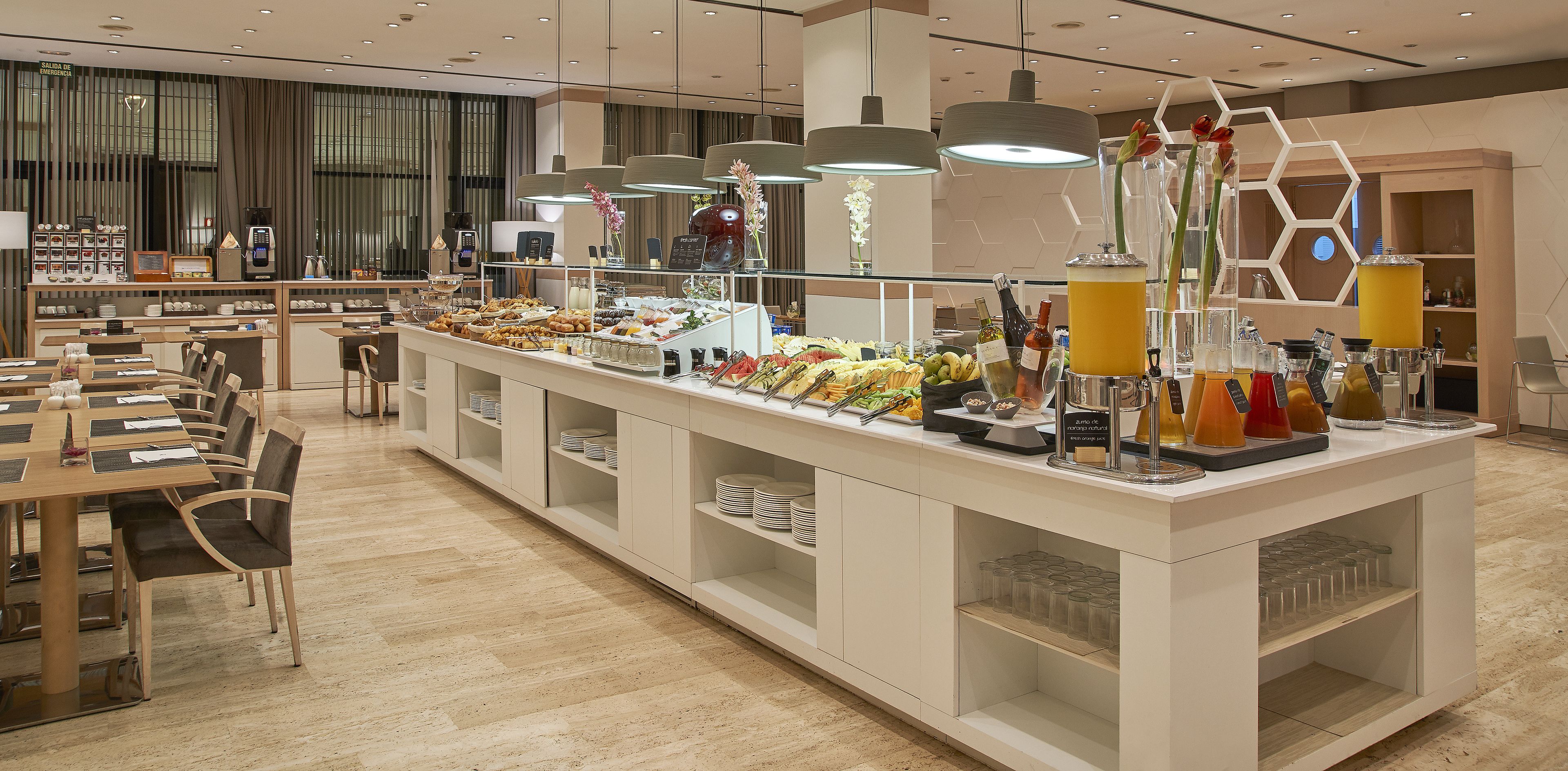 A buffet area with various food items and beverages, a seating area with tables and chairs, and a modern interior design with pendant lighting.