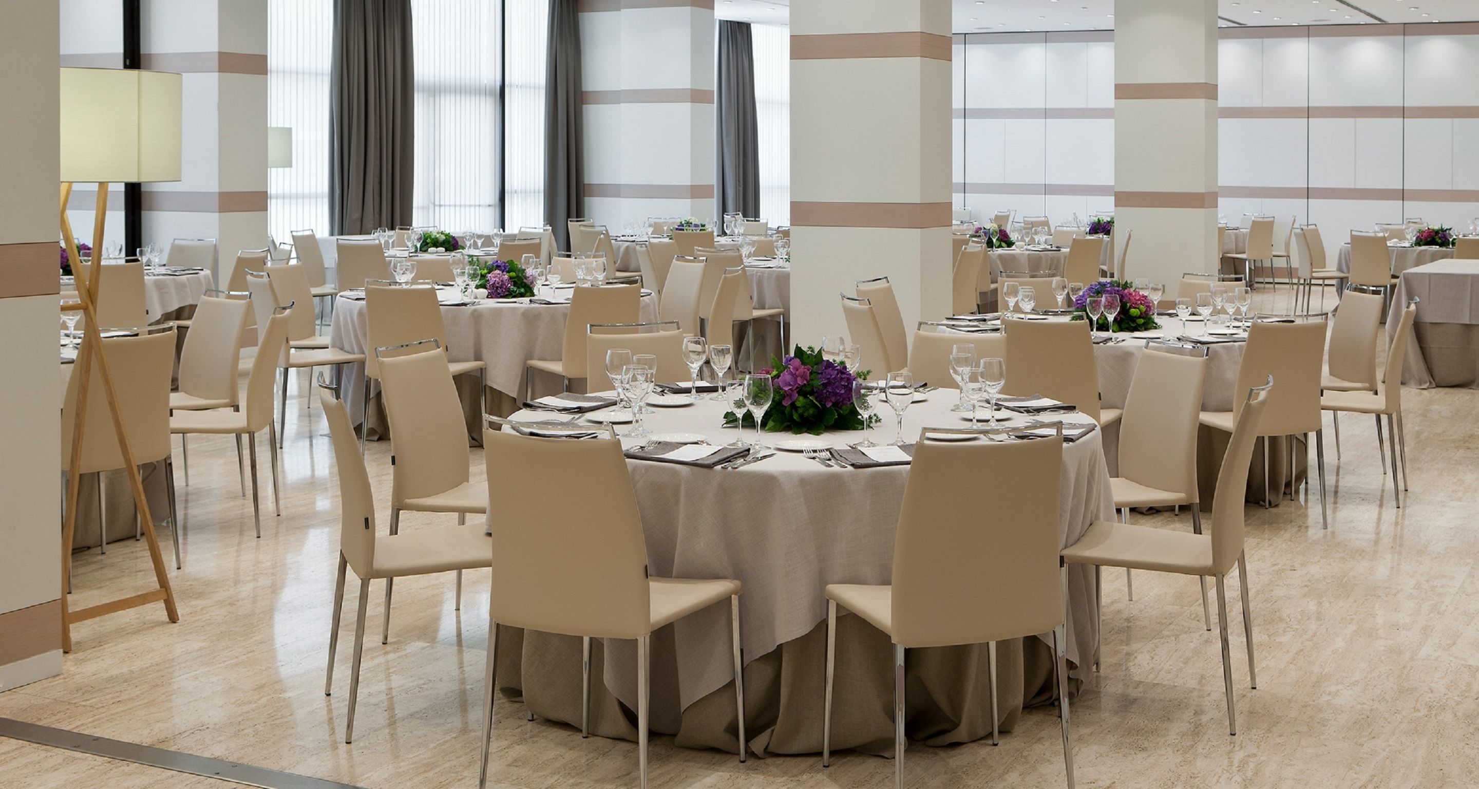 A banquet hall with round tables, beige chairs, and floral centerpieces.