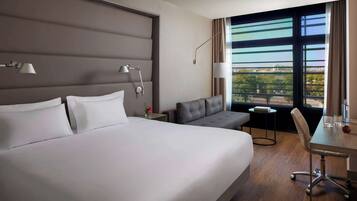 Habitación de hotel con una cama grande, un sofá, una pequeña mesa y una silla. Hay dos lámparas en la pared y una ventana con vistas a edificios.