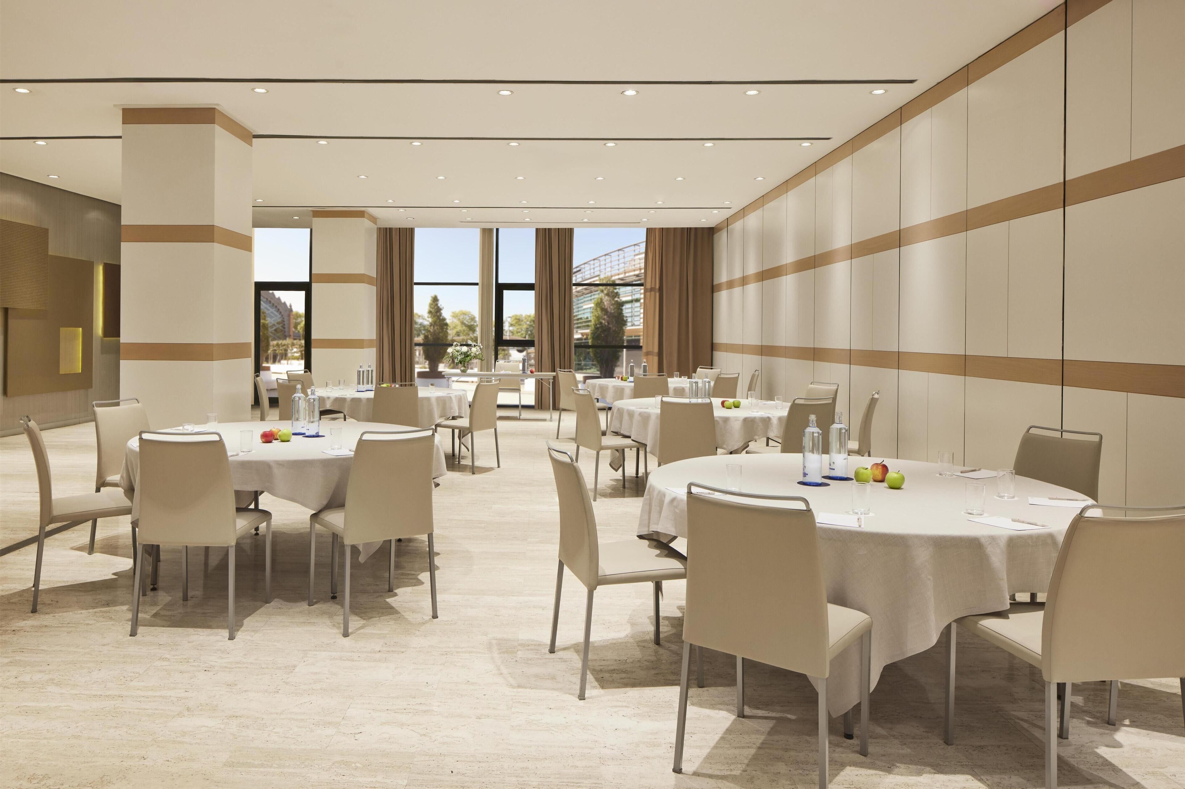 A spacious conference room with multiple round tables and chairs, large windows with curtains, and a modern interior design.