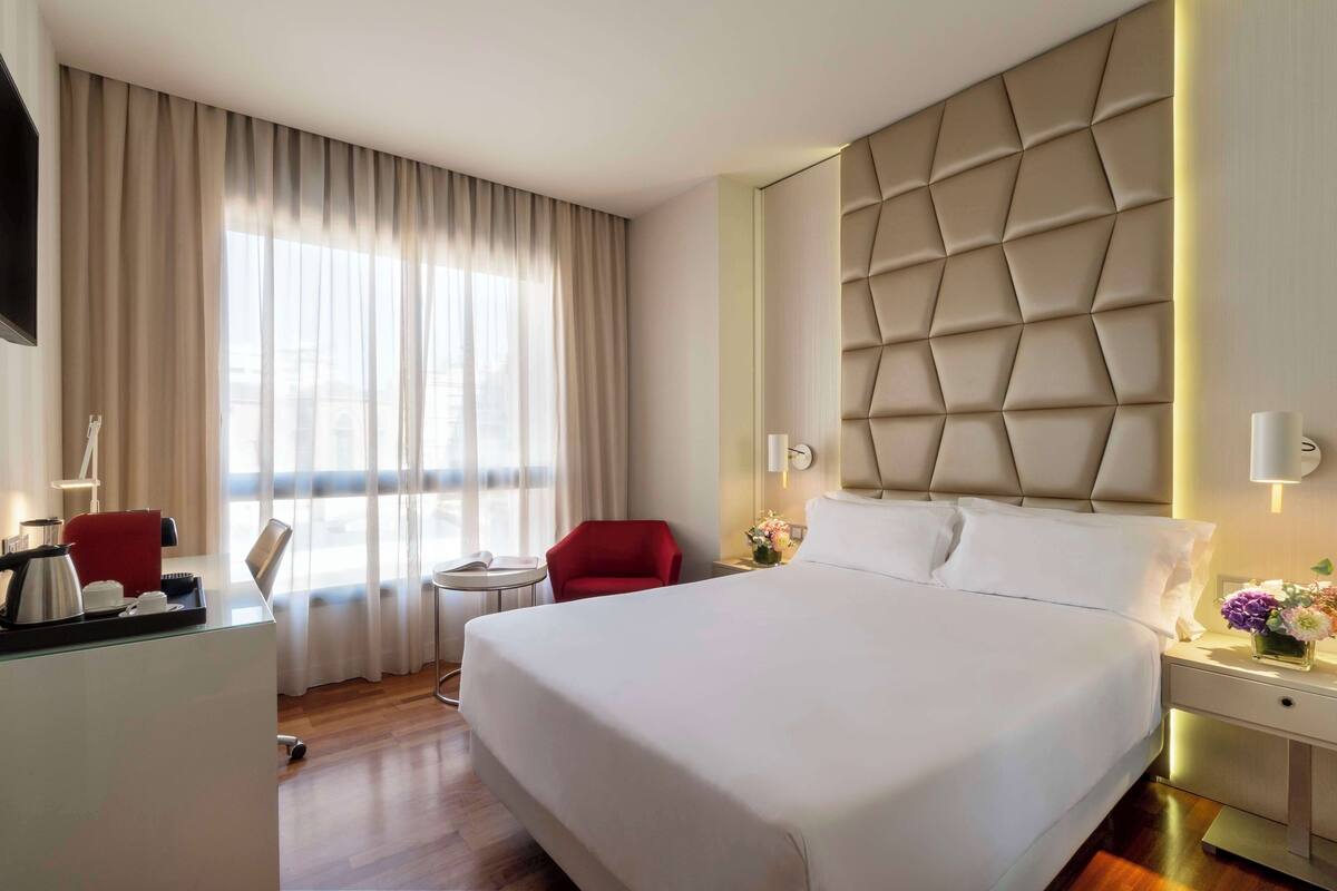 A hotel room with a large bed, a desk with a red chair, a small table, and a window with curtains.