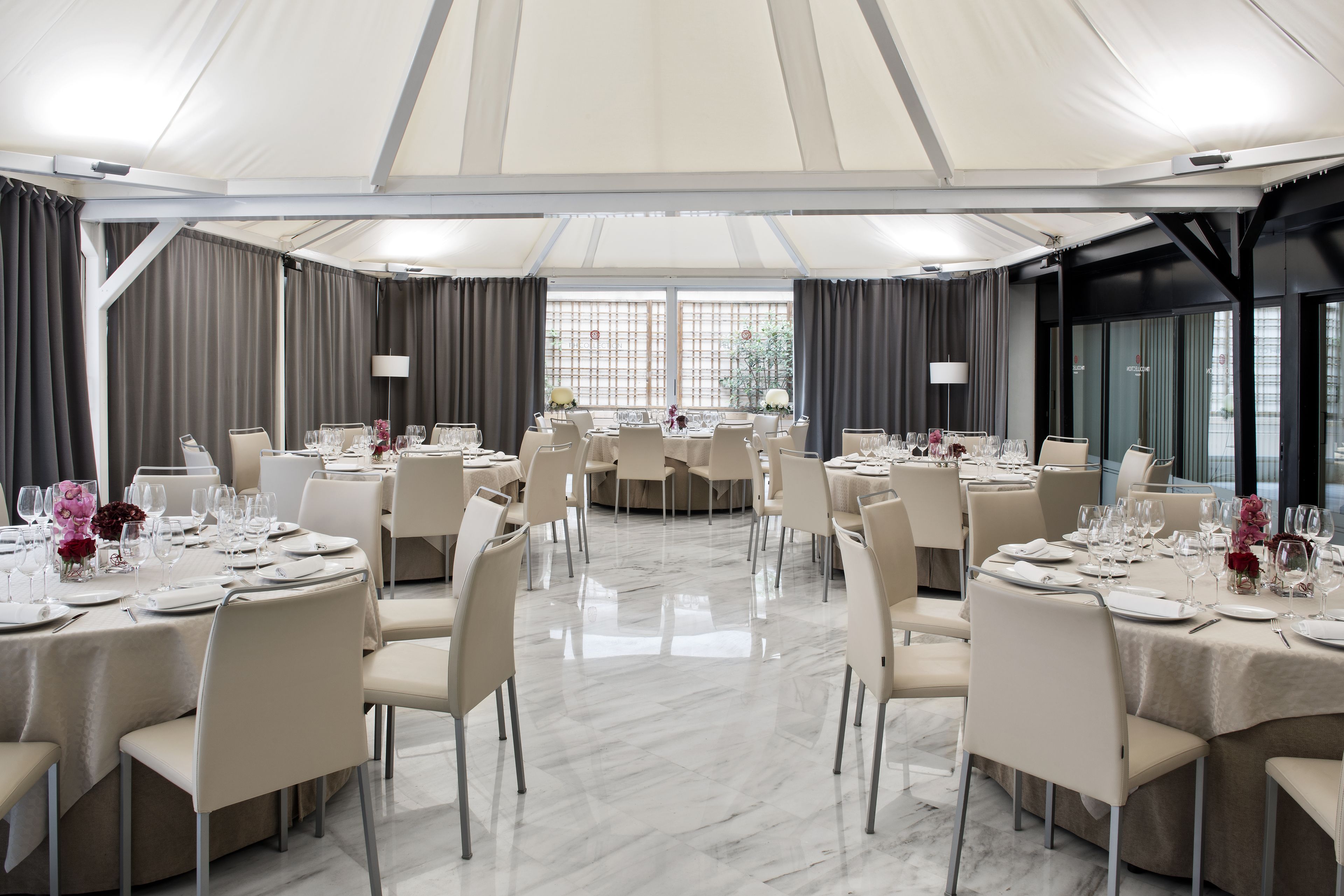 Une grande salle de banquet, bien éclairée, avec de nombreuses tables rondes dressées pour le repas, présentant un design moderne aux tons neutres et un sol en marbre.