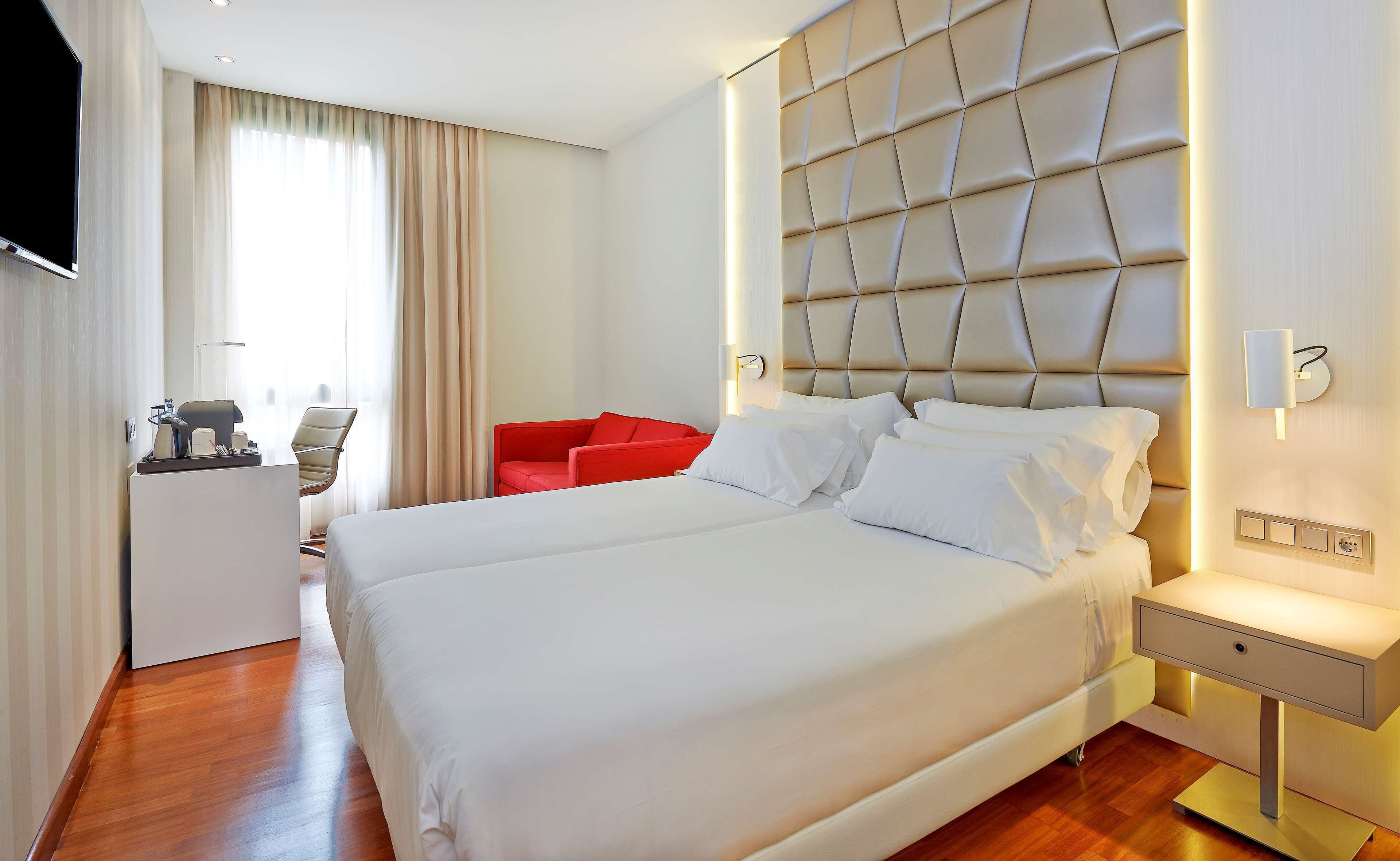 Habitación de hotel con una cama grande, una silla roja, un escritorio con lámpara y un televisor montado en la pared.