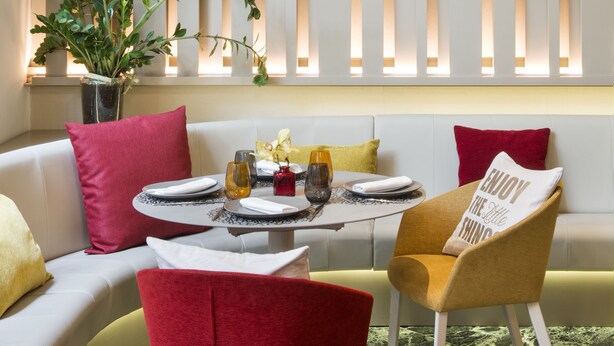Ein moderner Essbereich mit einem weißen Sofa, roten und gelben Stühlen, einem runden Tisch mit einer Vase sowie einer Wand mit vertikalen Lamellen und hängenden Lampen.