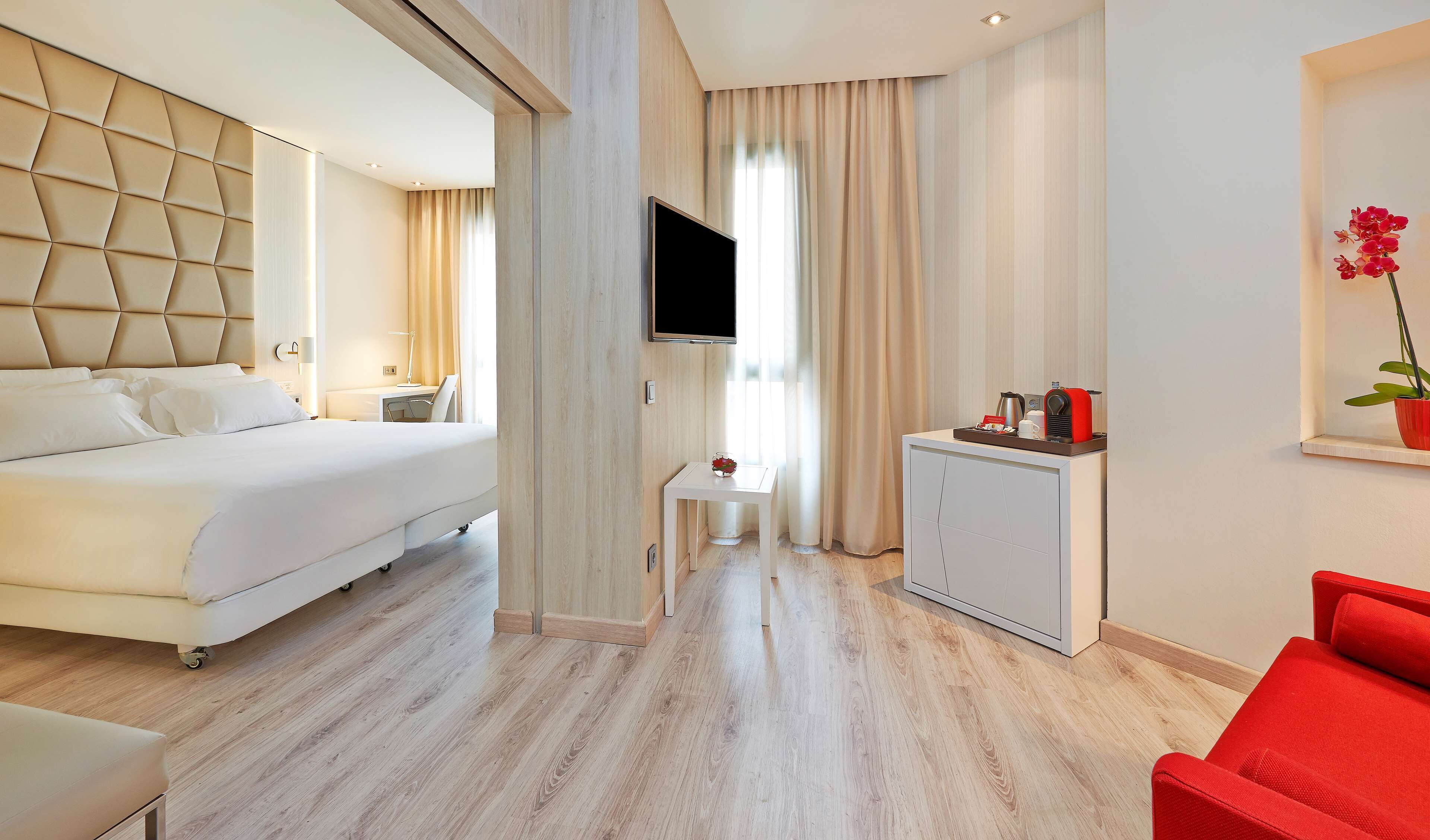 Une chambre d’hôtel moderne équipée d’un lit, d’un bureau, d’une télévision, d’une petite table ornée d’une fleur rouge et d’une chaise rouge.
