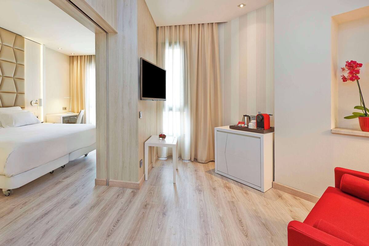 A hotel room with a bed, a desk, a TV, a small table, a red chair, and a cabinet.