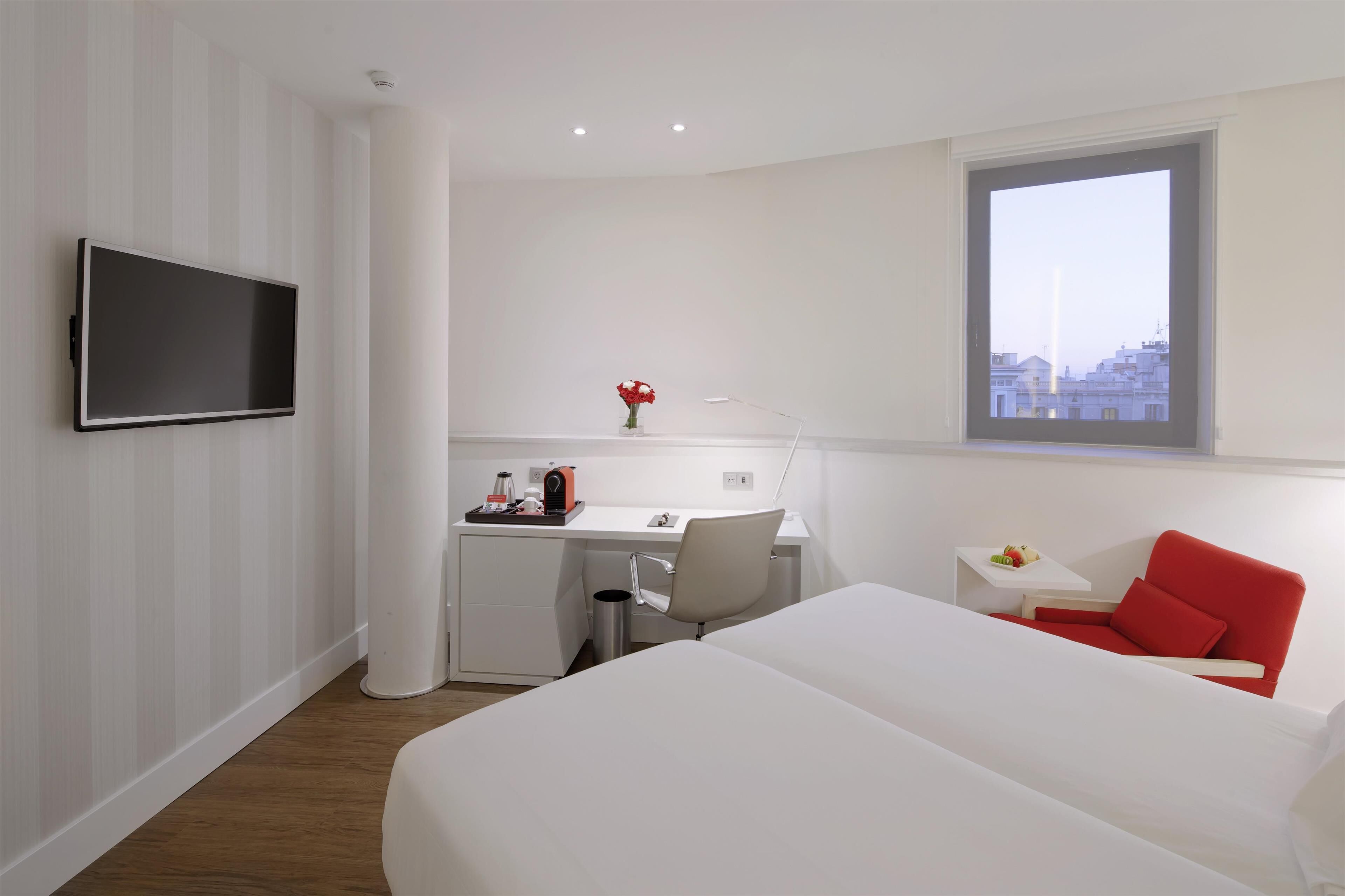 A hotel room with a bed, a desk with a chair, a TV mounted on the wall, and a window with a city view.