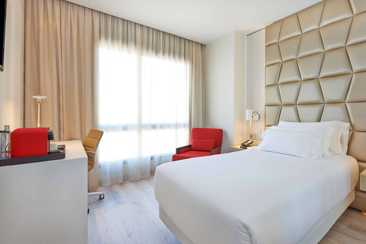 A hotel room with a large bed, a red armchair, a desk with a lamp, and a window with curtains.