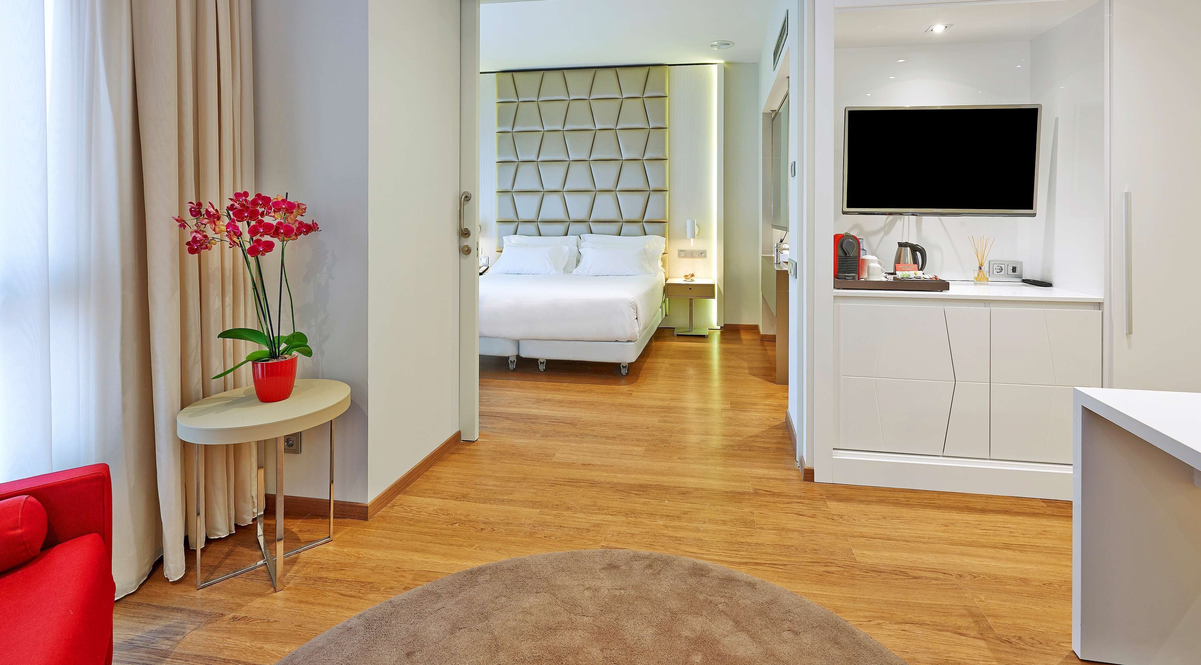 A hotel room with a bed, a TV, a small table with a red pot of flowers, and a red chair.
