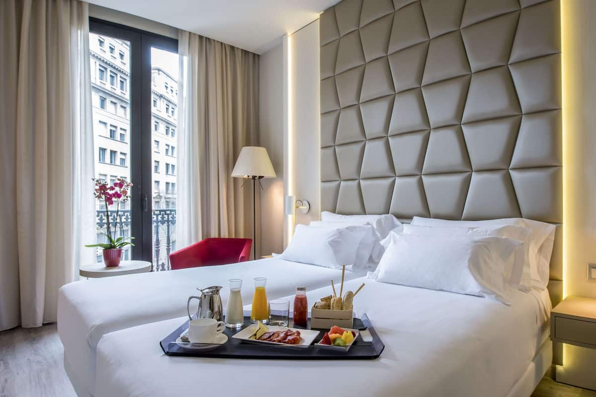 Une chambre d’hôtel avec un grand lit, un plateau avec des articles pour le petit-déjeuner, une chaise rouge et une vue sur les bâtiments par la fenêtre.