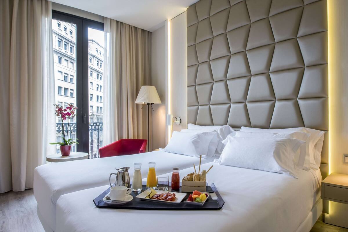 Habitación de hotel con una cama grande, una bandeja con alimentos para el desayuno, una silla roja y vistas a edificios por la ventana.