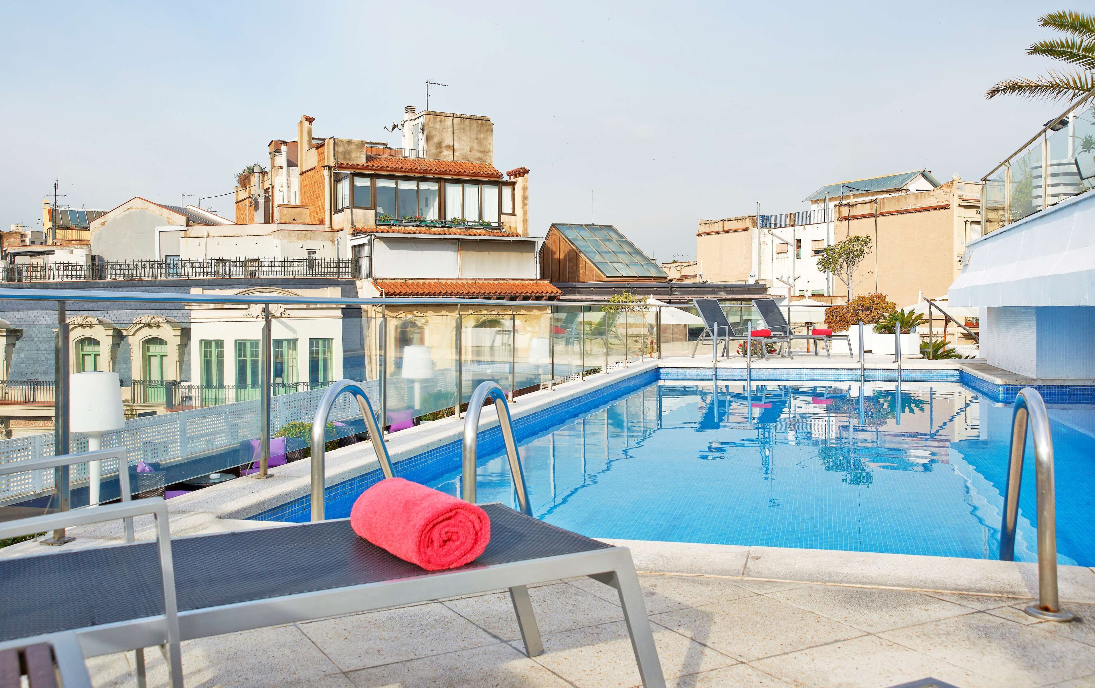 A rooftop pool with clear blue water, a red towel on a chair, and a view of surrounding buildings.