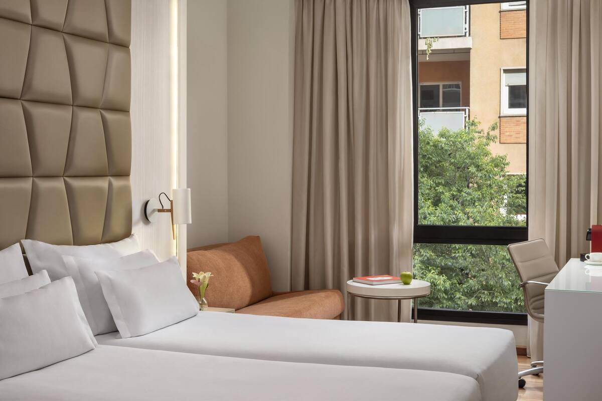 A hotel room with a bed, a small sofa, a round table with a book and a cup, and a view of greenery through the window.