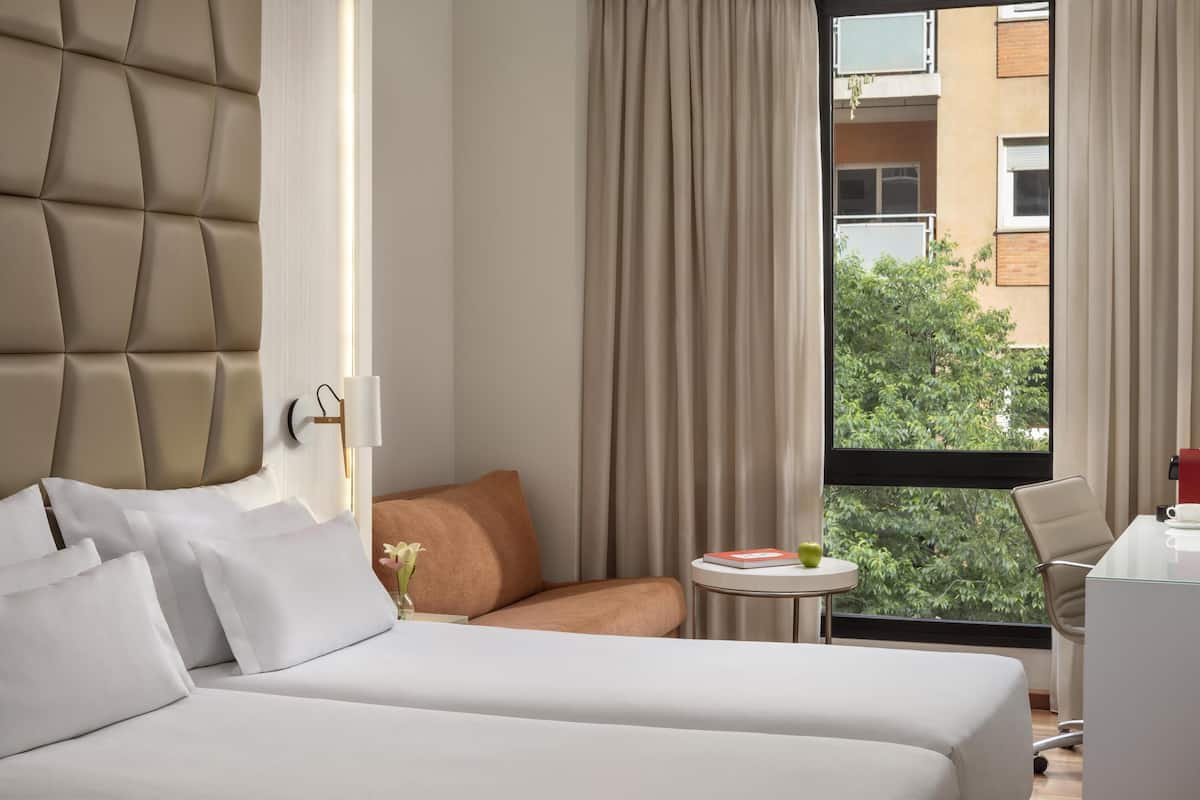 Habitación de hotel con cama, un pequeño sofá, una mesa redonda con un libro y una taza, y vista a zonas verdes por la ventana.