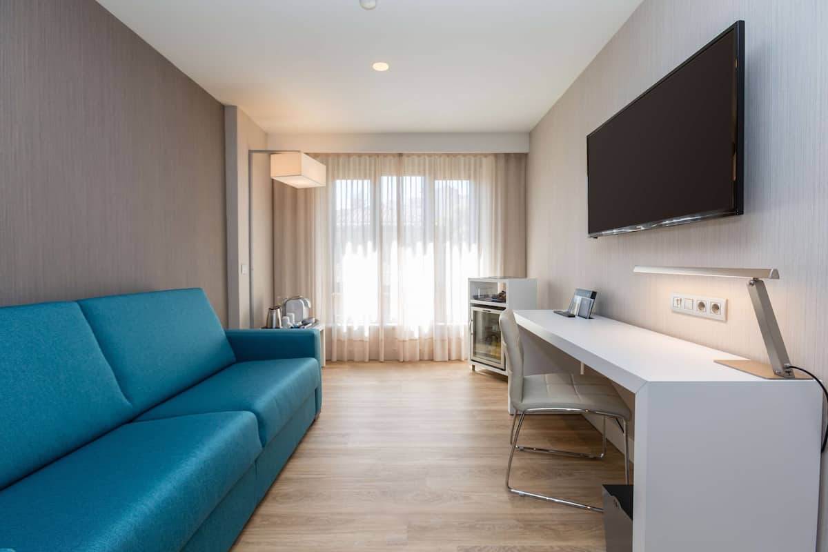 Ein modernes Hotelzimmer mit blauem Sofa, weißem Schreibtisch, Flachbildfernseher und einer Küchenzeile inklusive Kühlschrank und Backofen.