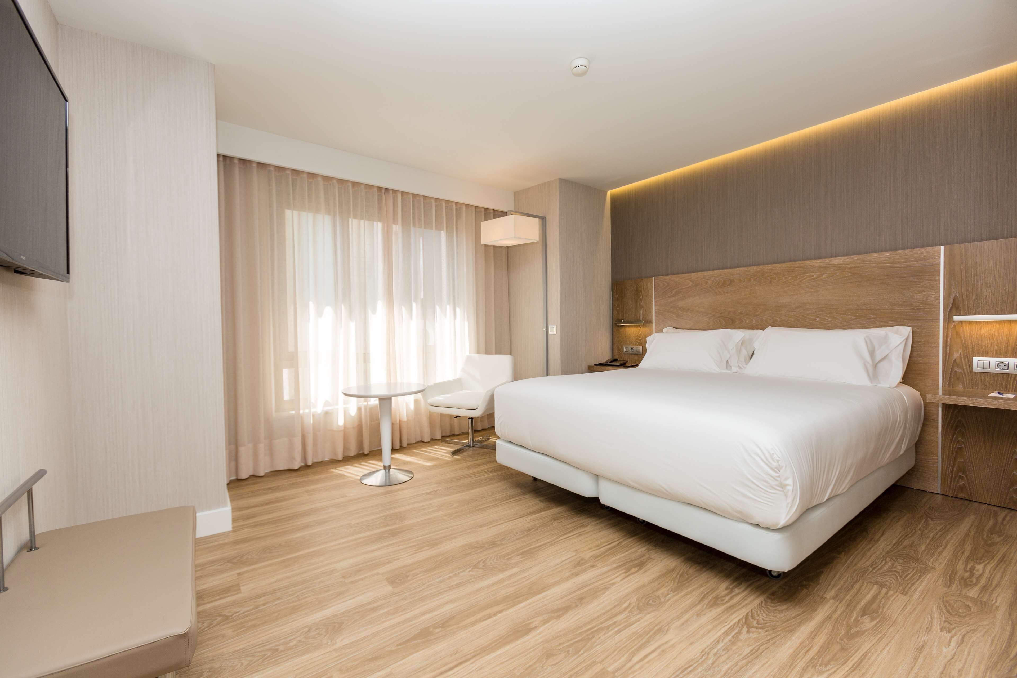 A modern hotel room with a large bed, a flat-screen TV, a bedside table, and a small round table.