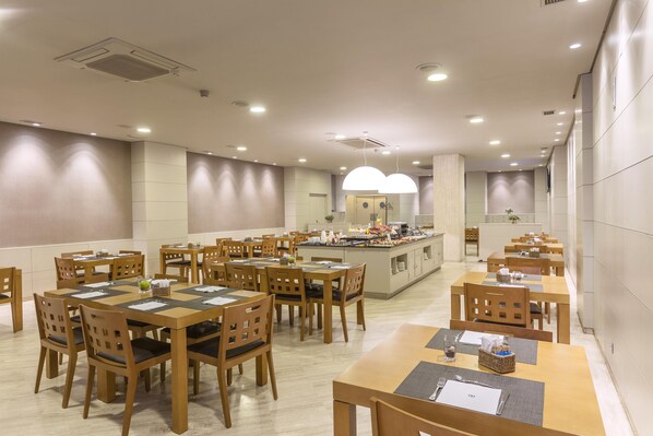 A spacious dining area with multiple wooden tables and chairs, a central island with a buffet, and recessed ceiling lighting. - NH Oviedo Principado (Oviedo)