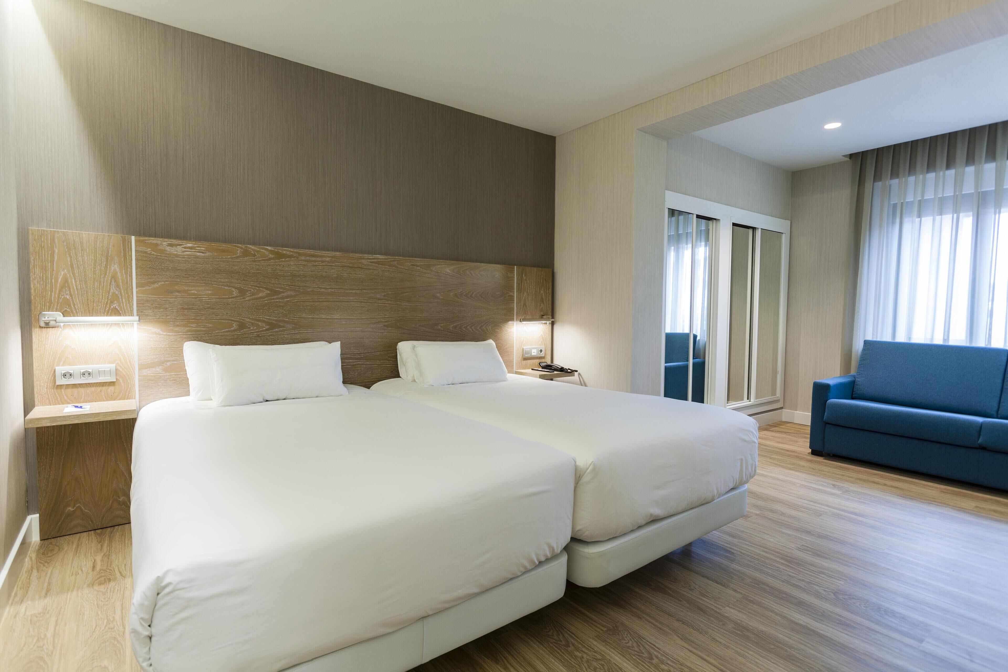 Habitación de hotel con dos camas, un sofá azul, suelo de madera y un ventanal con cortinas.