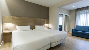 A hotel room with two beds, a blue sofa, wooden flooring, and a large window with curtains.