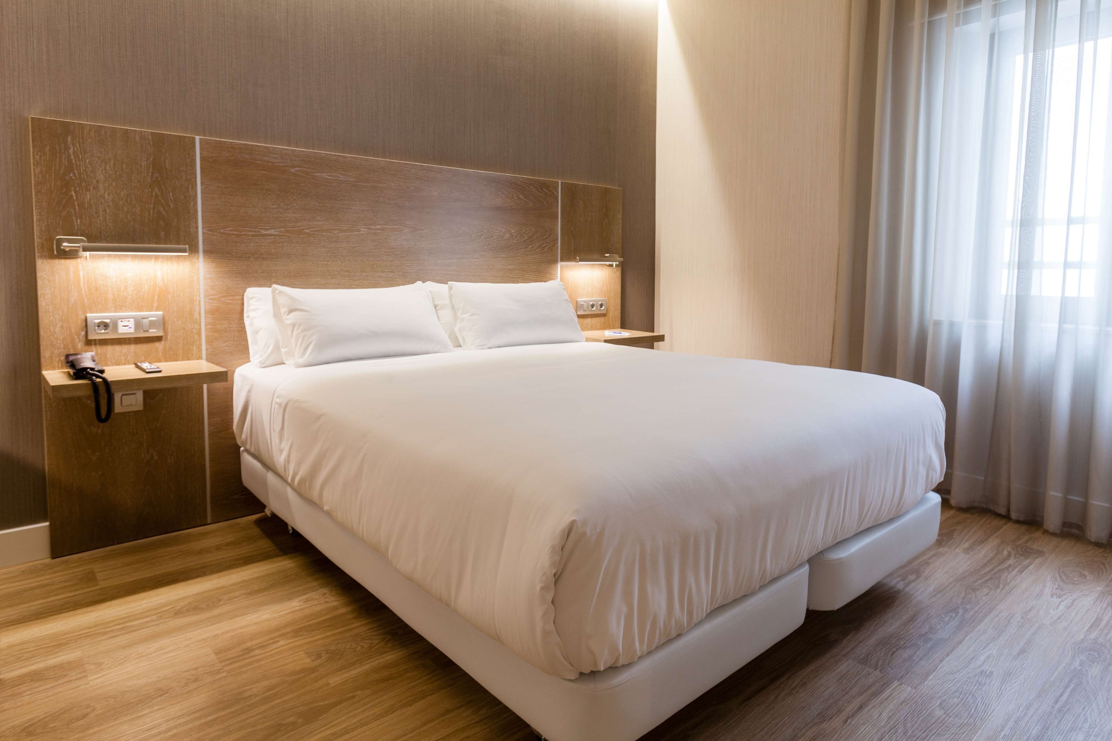 A modern hotel room with a large bed, wooden headboard, bedside lamps, and a power outlet on the nightstand.