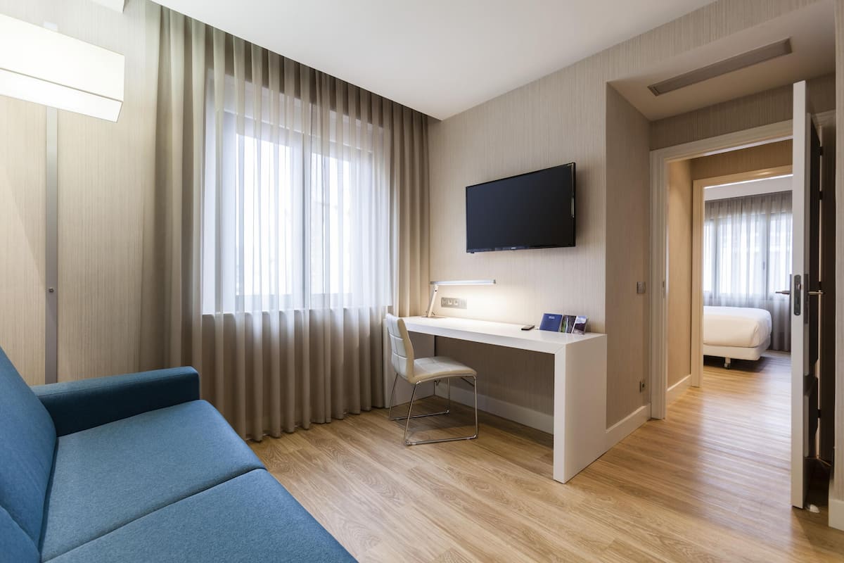 Ein modernes Hotelzimmer mit blauem Sofa, Schreibtisch mit Stuhl, Flachbildfernseher und Bett.