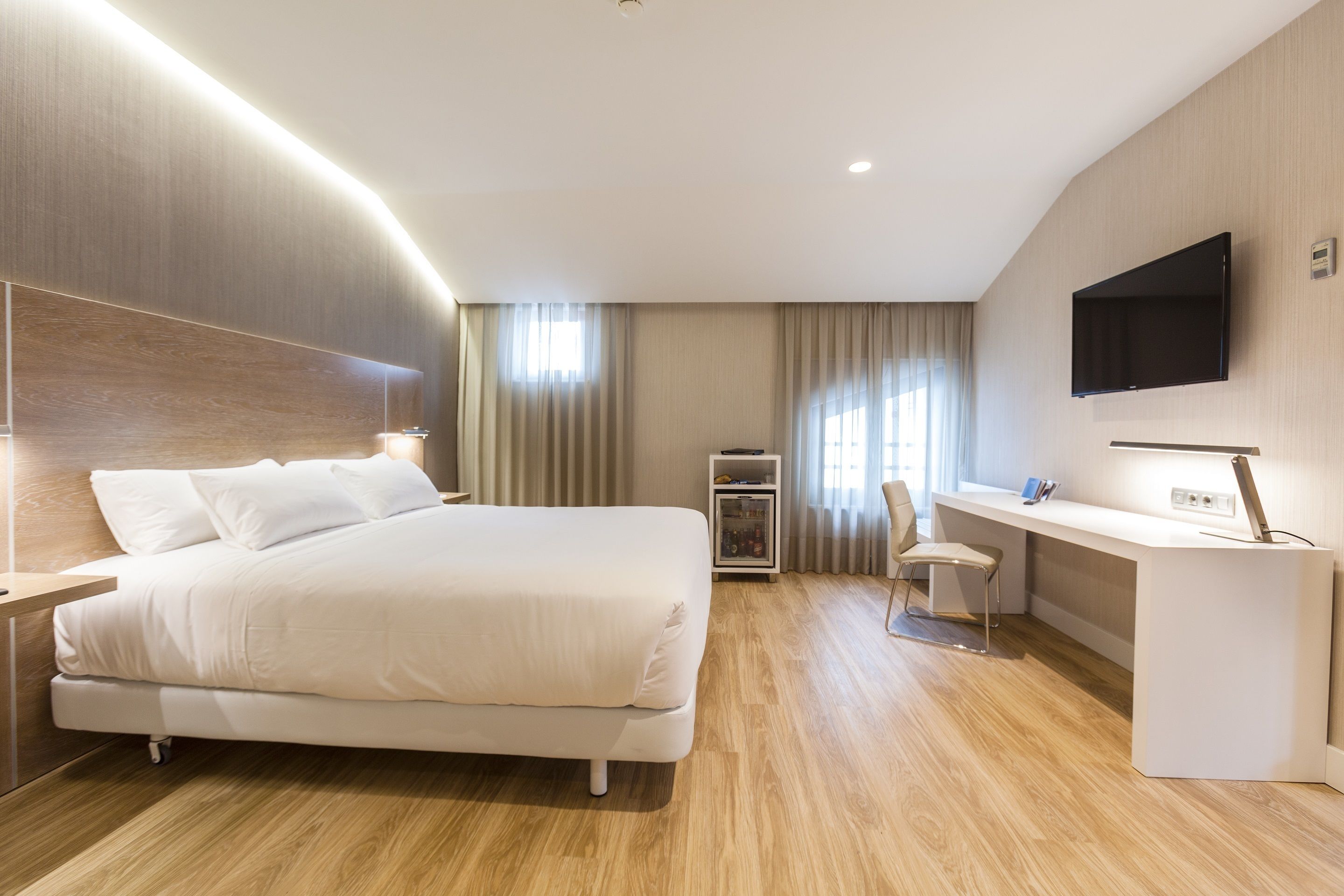 A modern hotel room with a large bed, a desk, a chair, and a flat-screen TV mounted on the wall.
