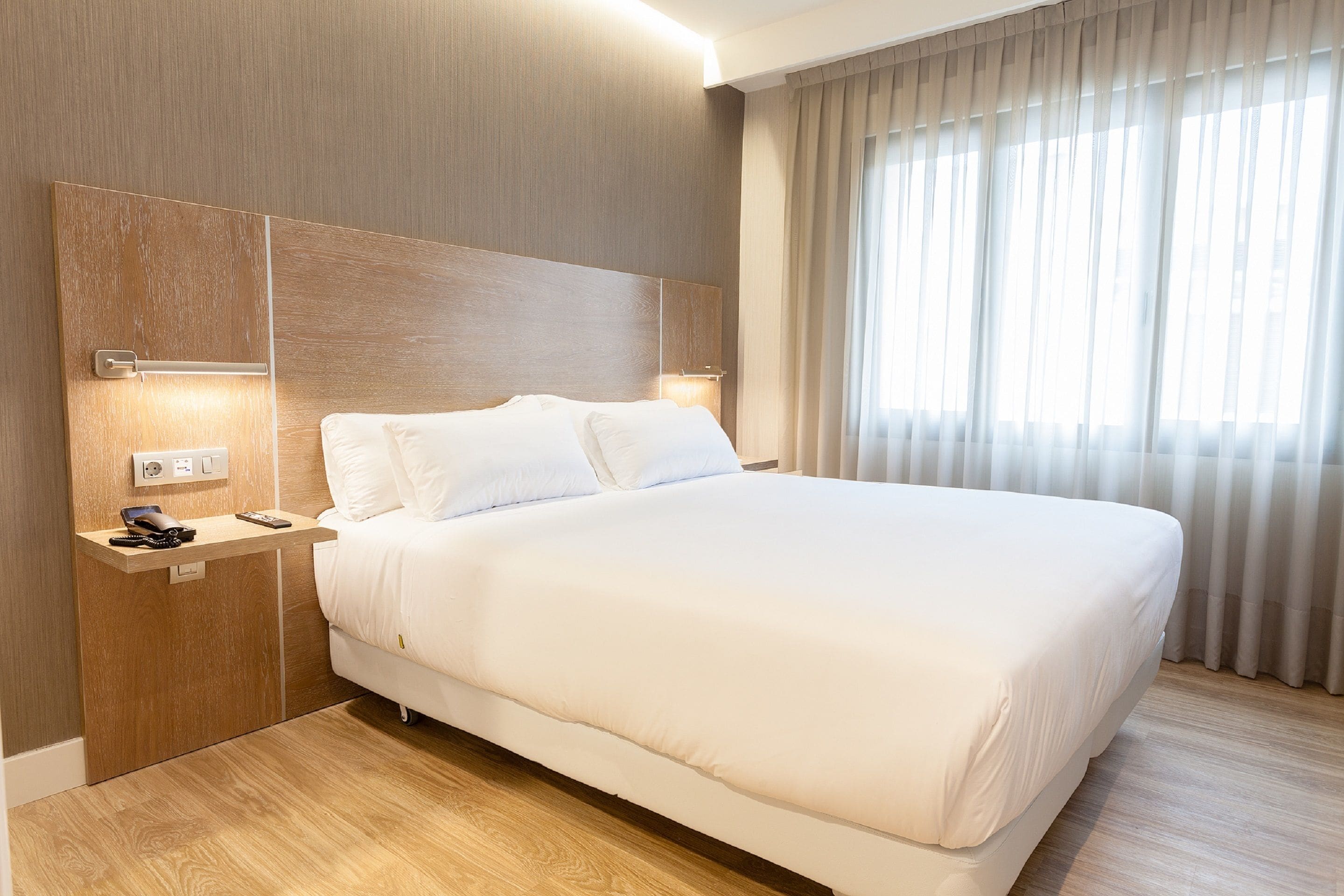 Een moderne hotelkamer met een groot bed, een houten hoofdbord, nachtkastjes met lampen en een groot raam met gordijnen.
