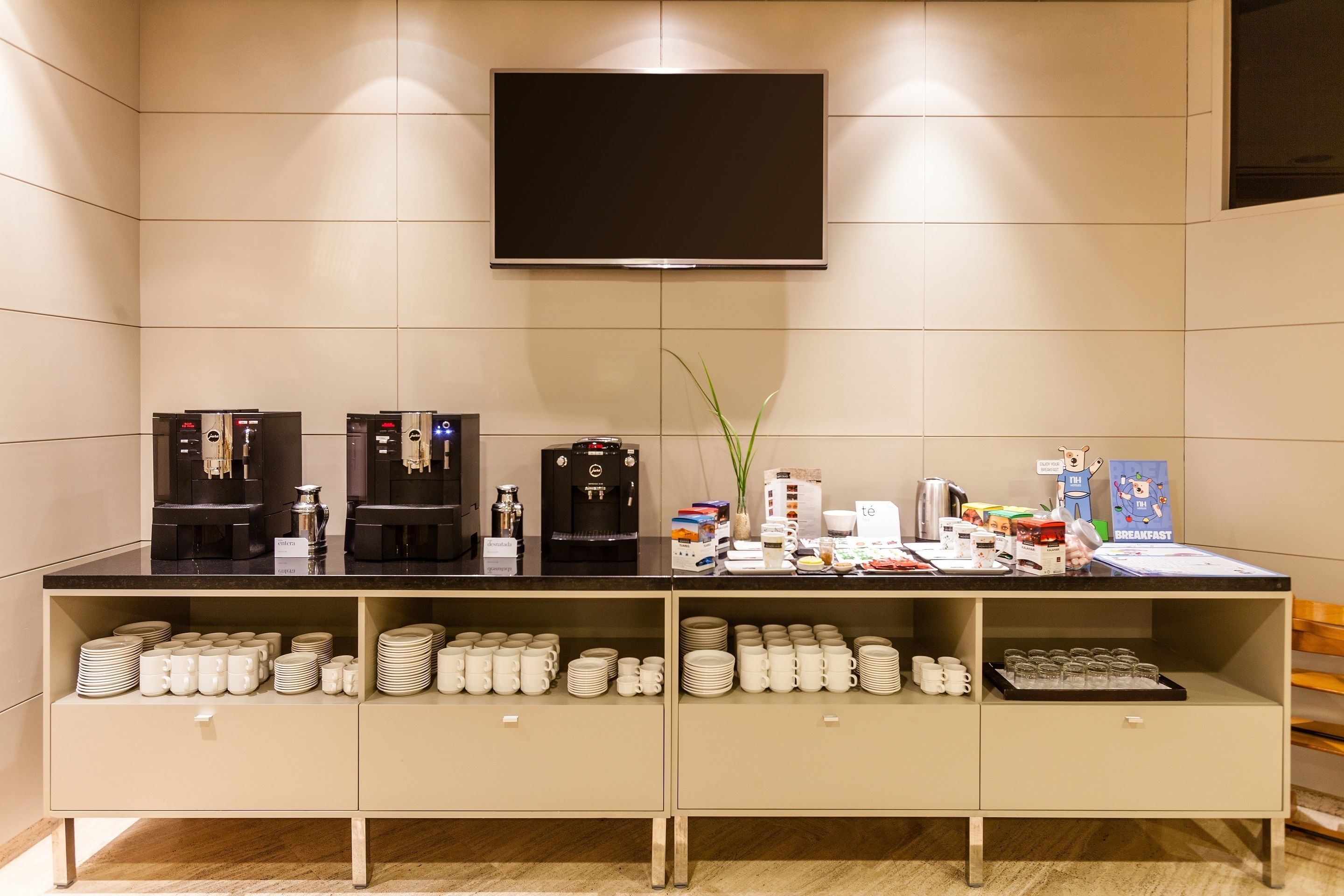 Una barra de desayuno con máquinas de café, un televisor de pantalla plana y tazas y platos ordenados.