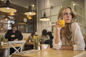 A person sitting at a table in a restaurant with hanging lights, another person in the background, and a bottle of olive oil on the table.