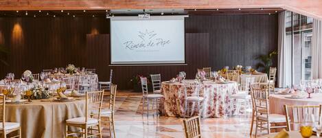 Un salón de banquetes con varias mesas redondas dispuestas para un evento formal, con centros de mesa florales y sillas con detalles dorados.