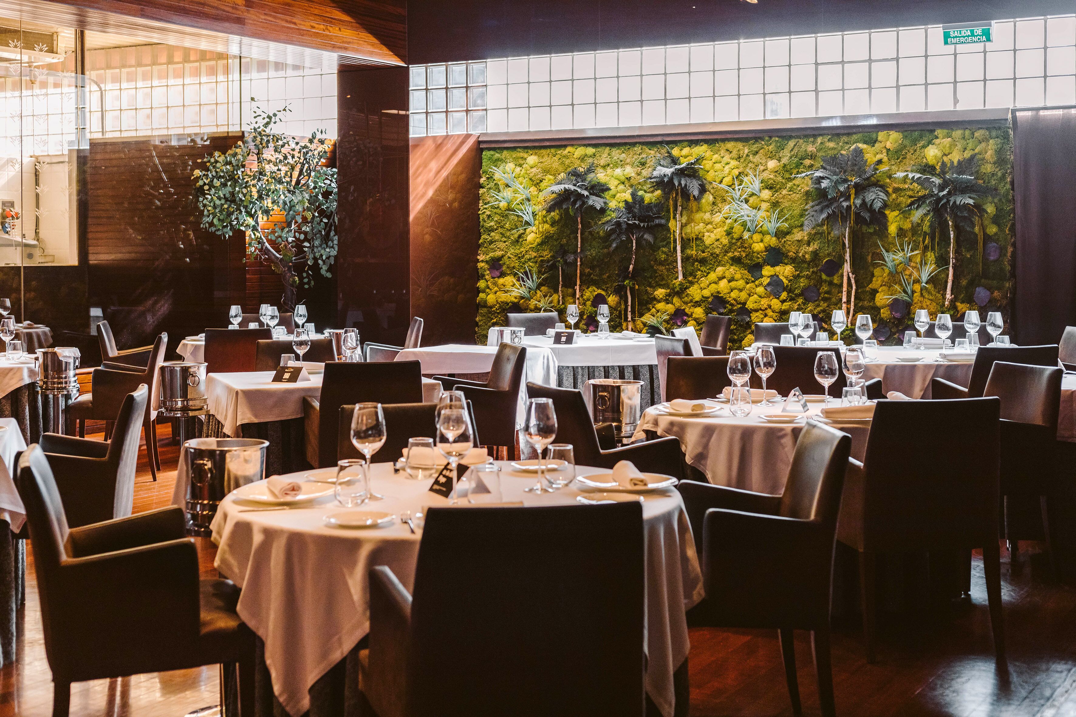 A restaurant with multiple tables set for dining, a large wall with a tropical plant display, and a visible emergency exit sign.