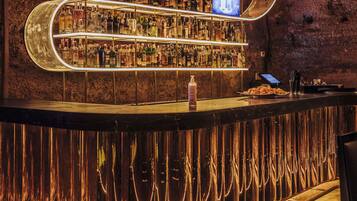 Un bar con estantes llenos de botellas de licor, una pantalla digital y una barra con una botella y vasos.
