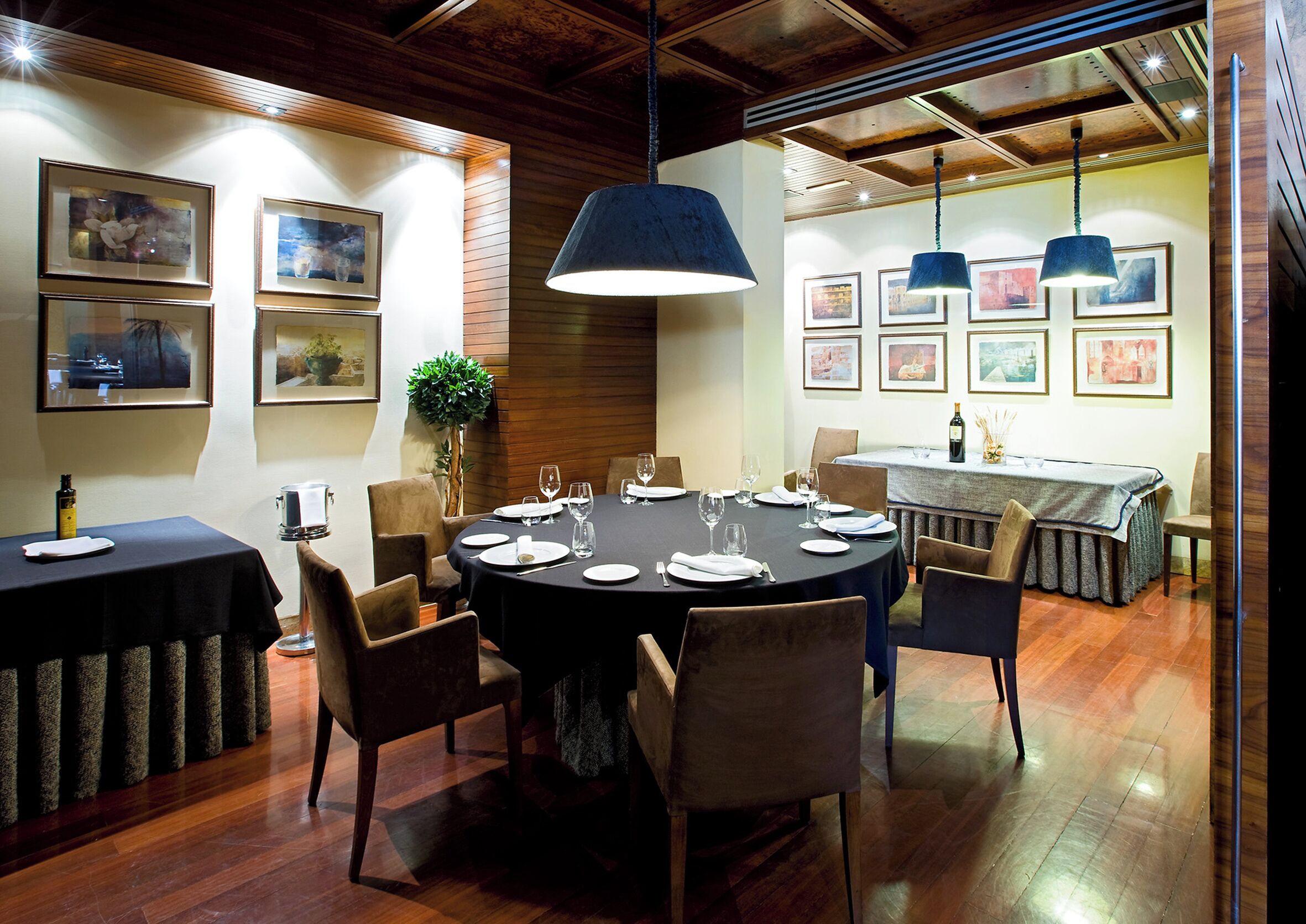 Un espace repas avec des tables rondes disposées pour accueillir six personnes, un plancher en bois et des œuvres d’art encadrées accrochées aux murs.