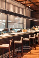 Un área de bar moderna con taburetes de madera, una vitrina de vidrio con alimentos y una variedad de botellas en la barra.