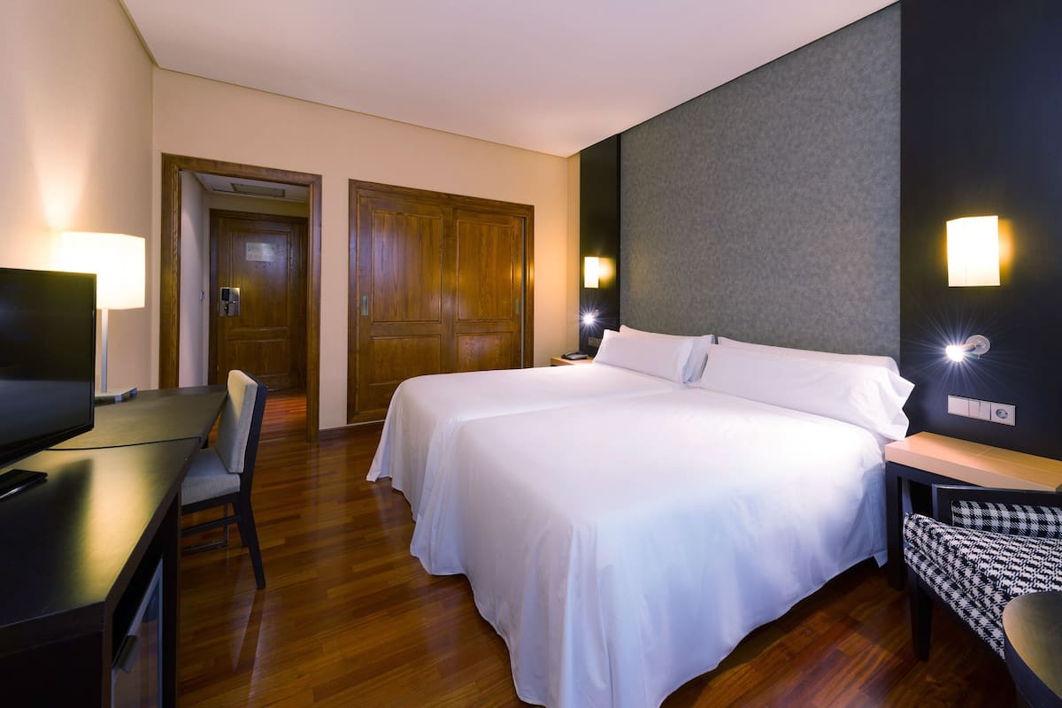 Une chambre d’hôtel avec deux lits, un bureau avec une chaise, une télévision et un luminaire fixé au mur.