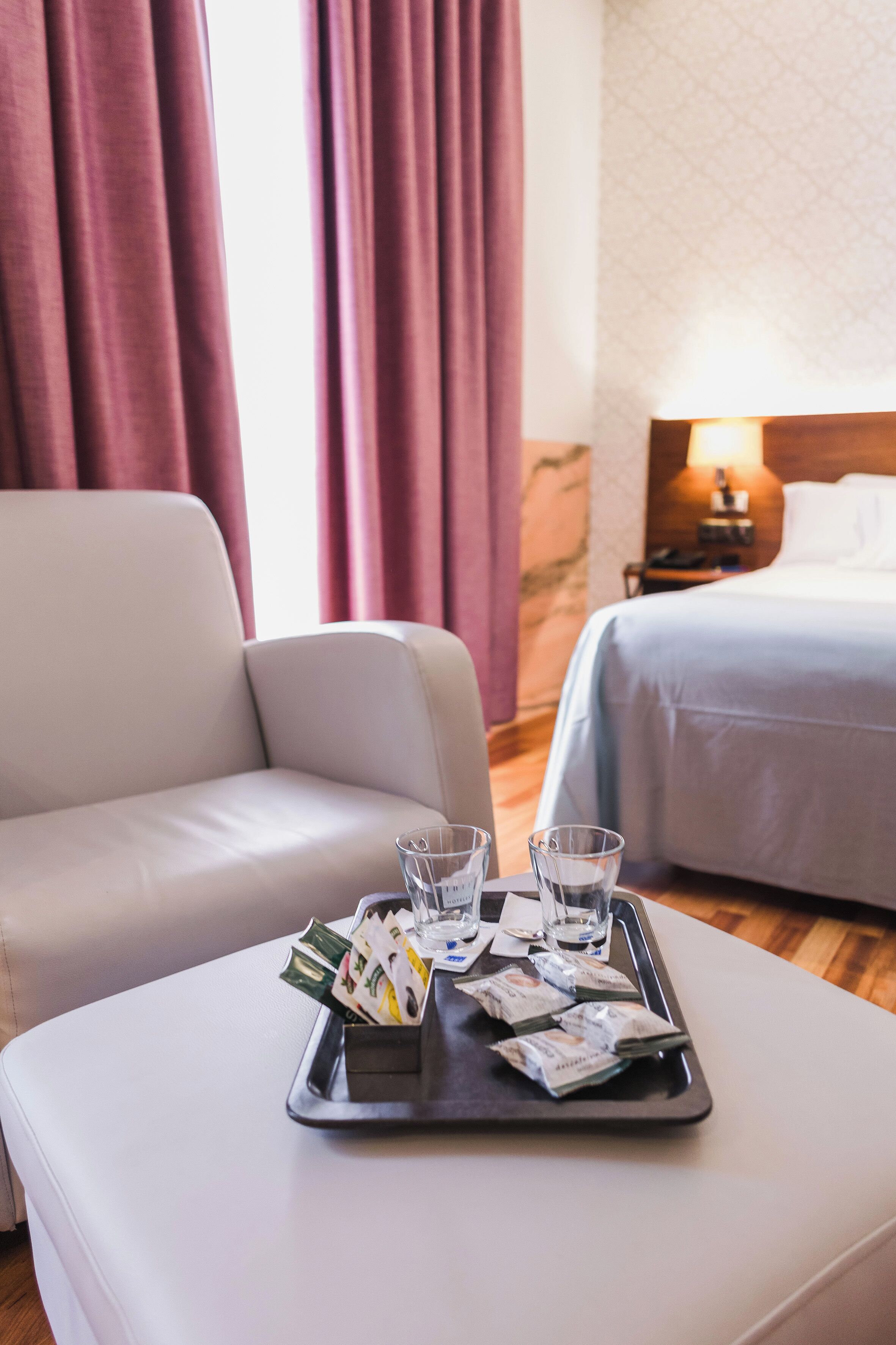 Ein Hotelzimmer mit Bett, einer Sitzecke, einem Tablett mit Gläsern und Geld sowie einem Nachttisch mit Lampe.