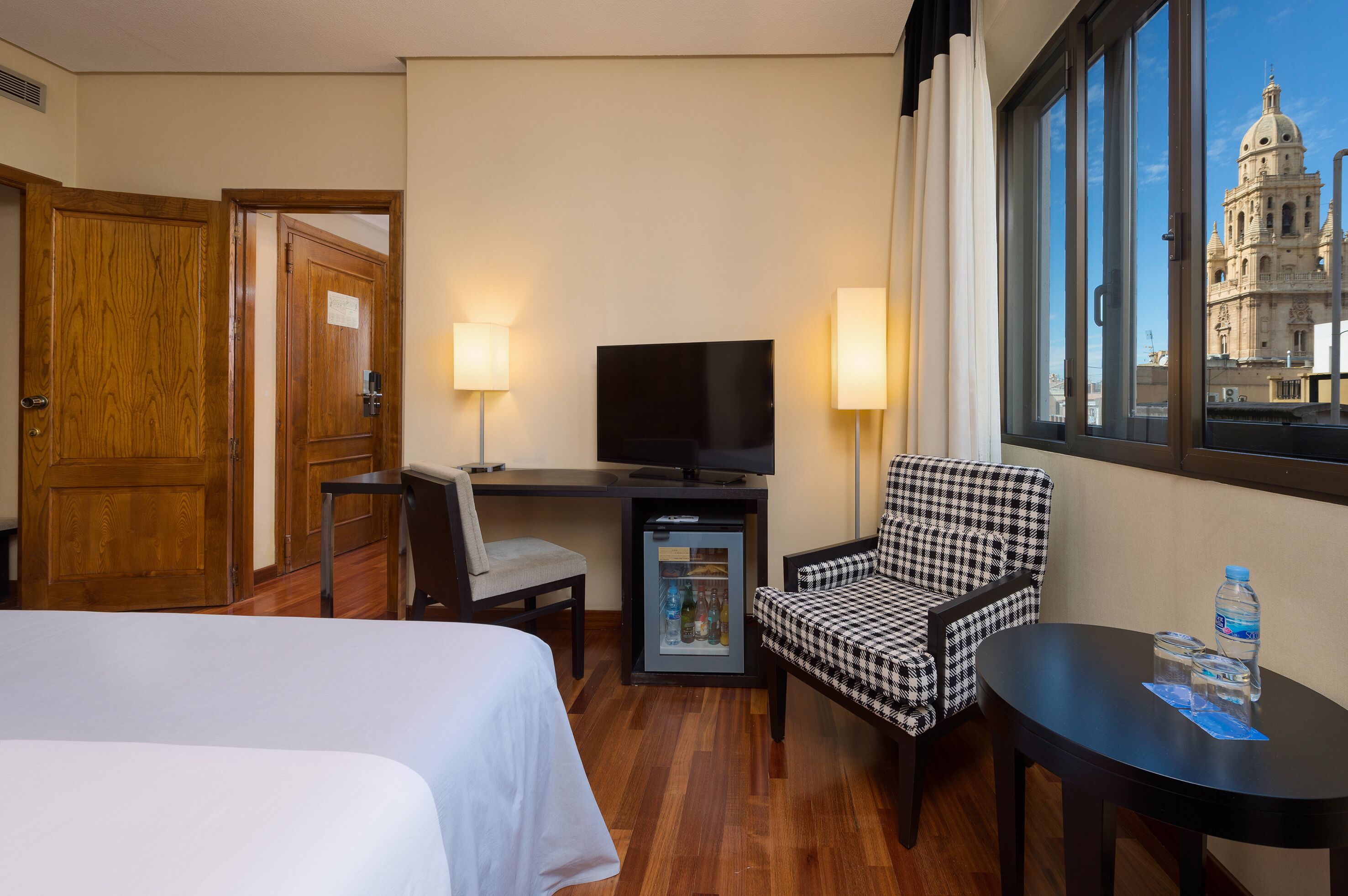 Habitación de hotel con cama, escritorio con televisor, silla, refrigerador pequeño, mesa con botellas de agua y ventana con vista a un edificio histórico.