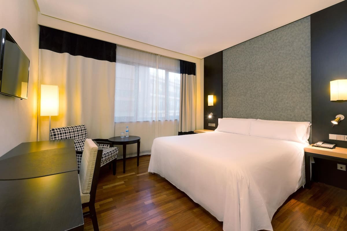 A hotel room with a bed, a desk, a chair, a TV, and a window with curtains.