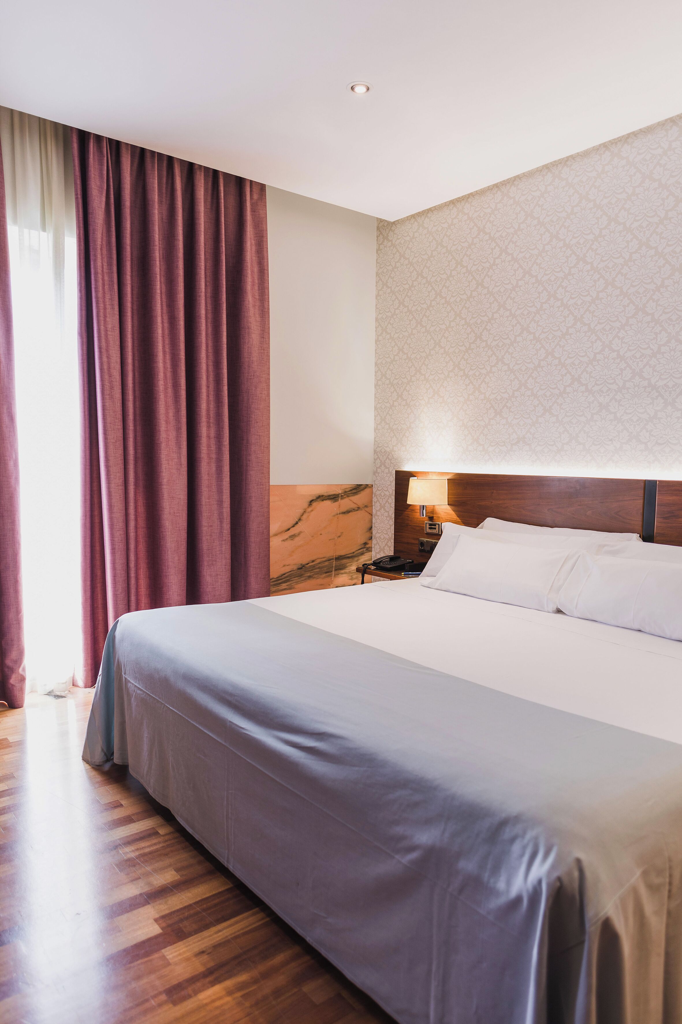 Una habitación de hotel con una cama grande, suelo de madera, una mesita de noche con lámpara y un papel tapiz estampado.