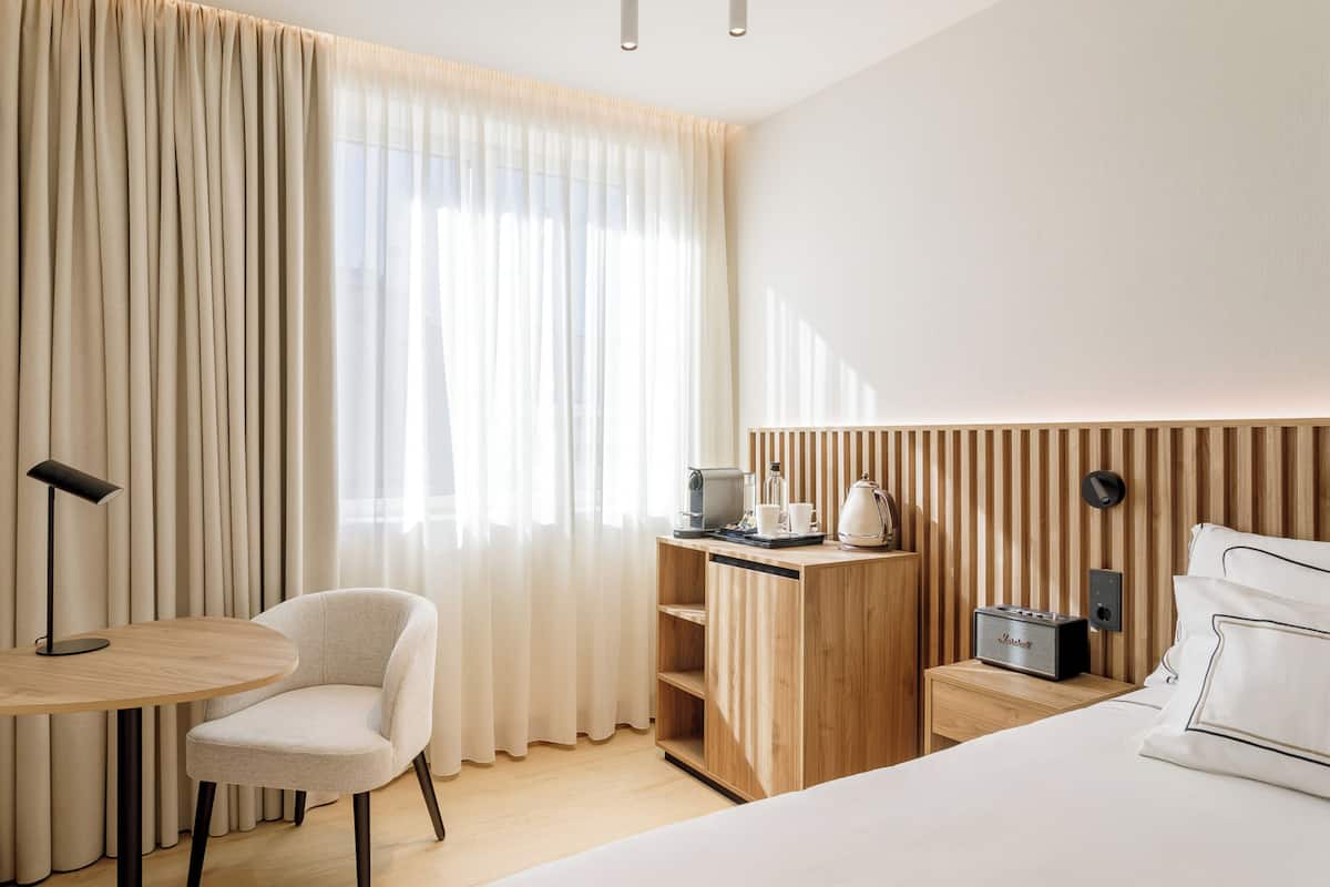Une chambre d’hôtel avec un lit, un bureau doté d’une radio, une chaise, une petite table et une fenêtre avec des rideaux.