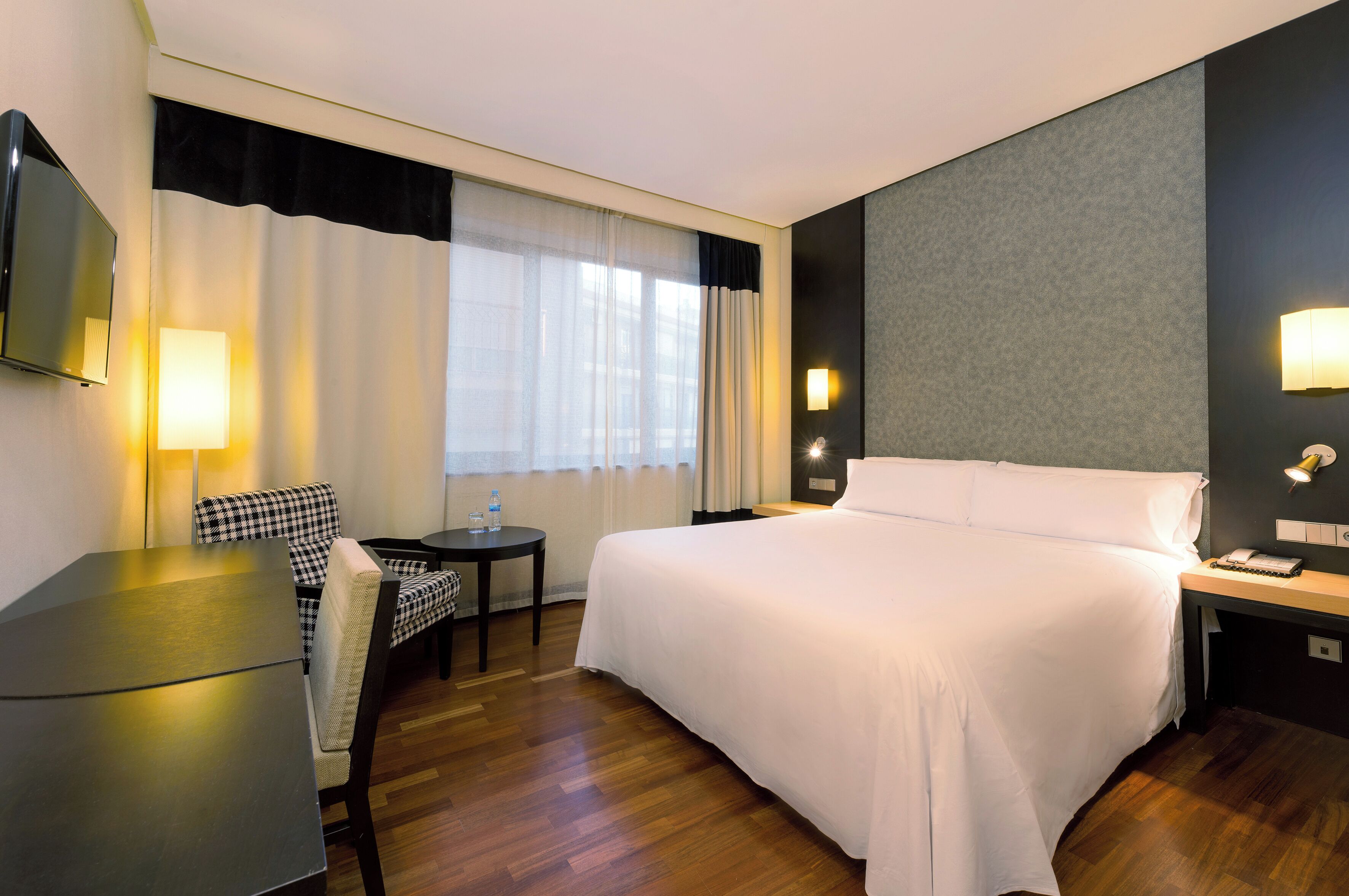 Habitación de hotel con una cama grande, un escritorio, una silla, un televisor y una ventana con cortinas.