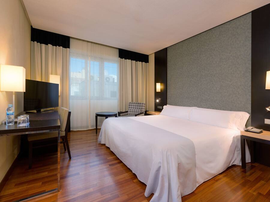A hotel room with a large bed, a desk with a chair, a television, and a window with curtains.