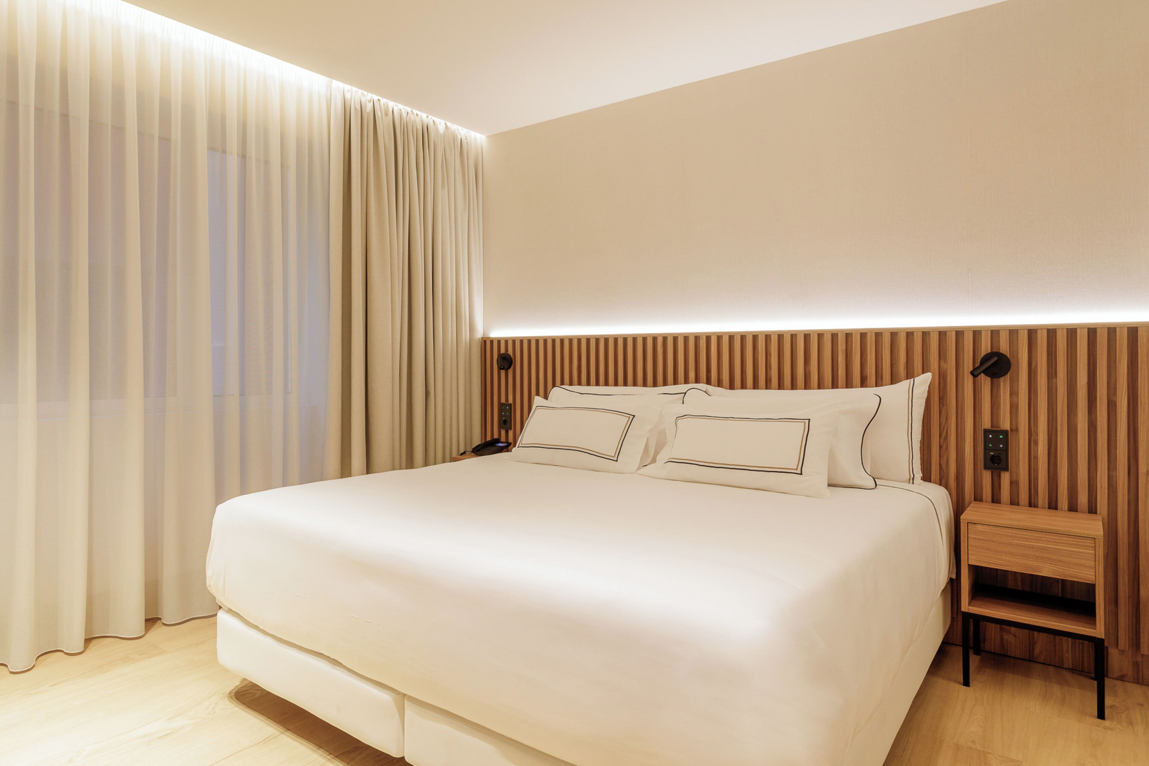 A modern hotel room with a large bed, a wooden headboard, a bedside table, and a wall-mounted control panel.