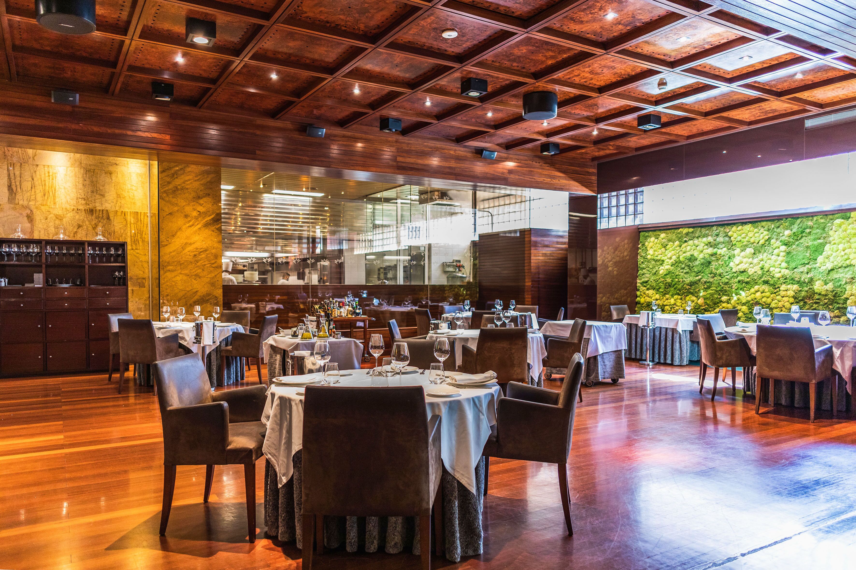 Un restaurante con suelo de madera, mesas dispuestas con manteles blancos y sillas, y una gran pared verde al fondo.